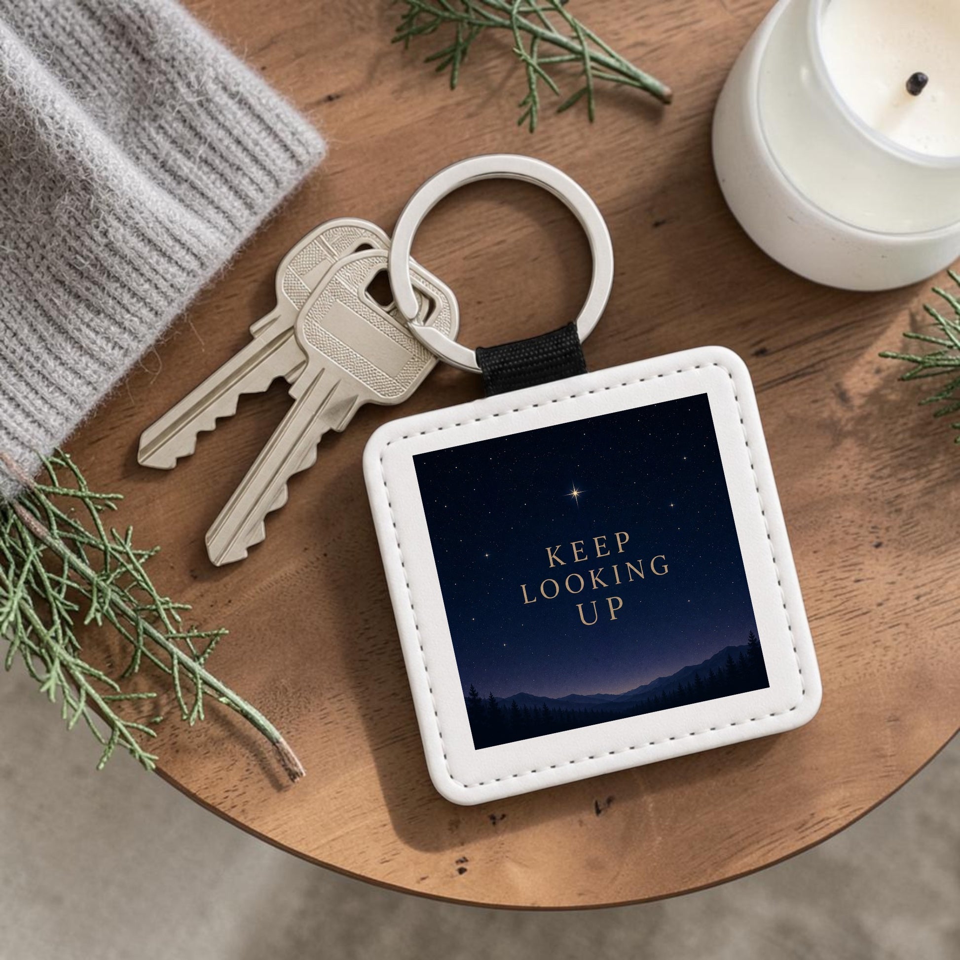The Keep Looking Up Celestial Keychain by Mirabilia Boutique—a square, inspirational accessory featuring the words “KEEP LOOKING UP” against a starry night sky—is shown beside two keys, a candle, a gray knit item, and green sprigs on a wooden surface.