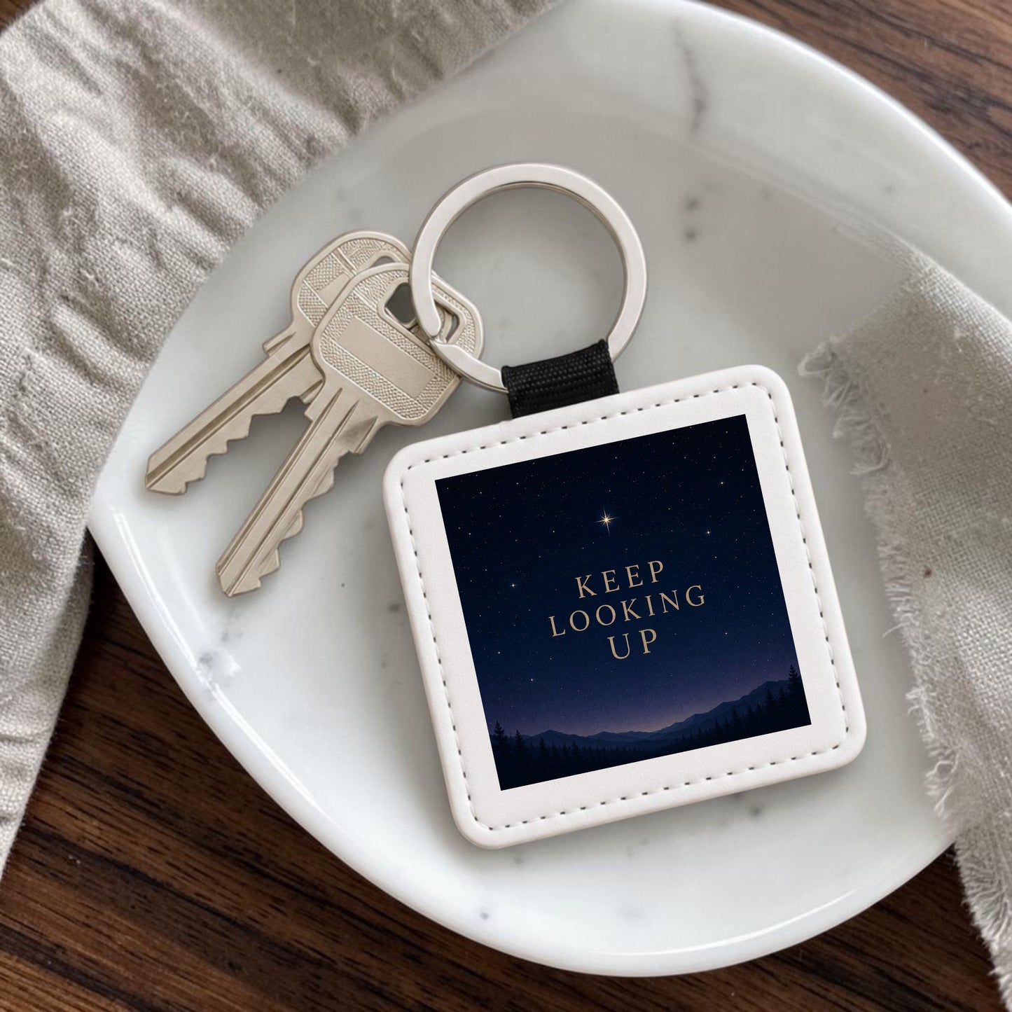 A Keep Looking Up Celestial Keychain by Mirabilia Boutique, featuring a starry night sky and the words “KEEP LOOKING UP,” is displayed with two silver keys on a white dish resting on a wooden surface beside a beige textured cloth.