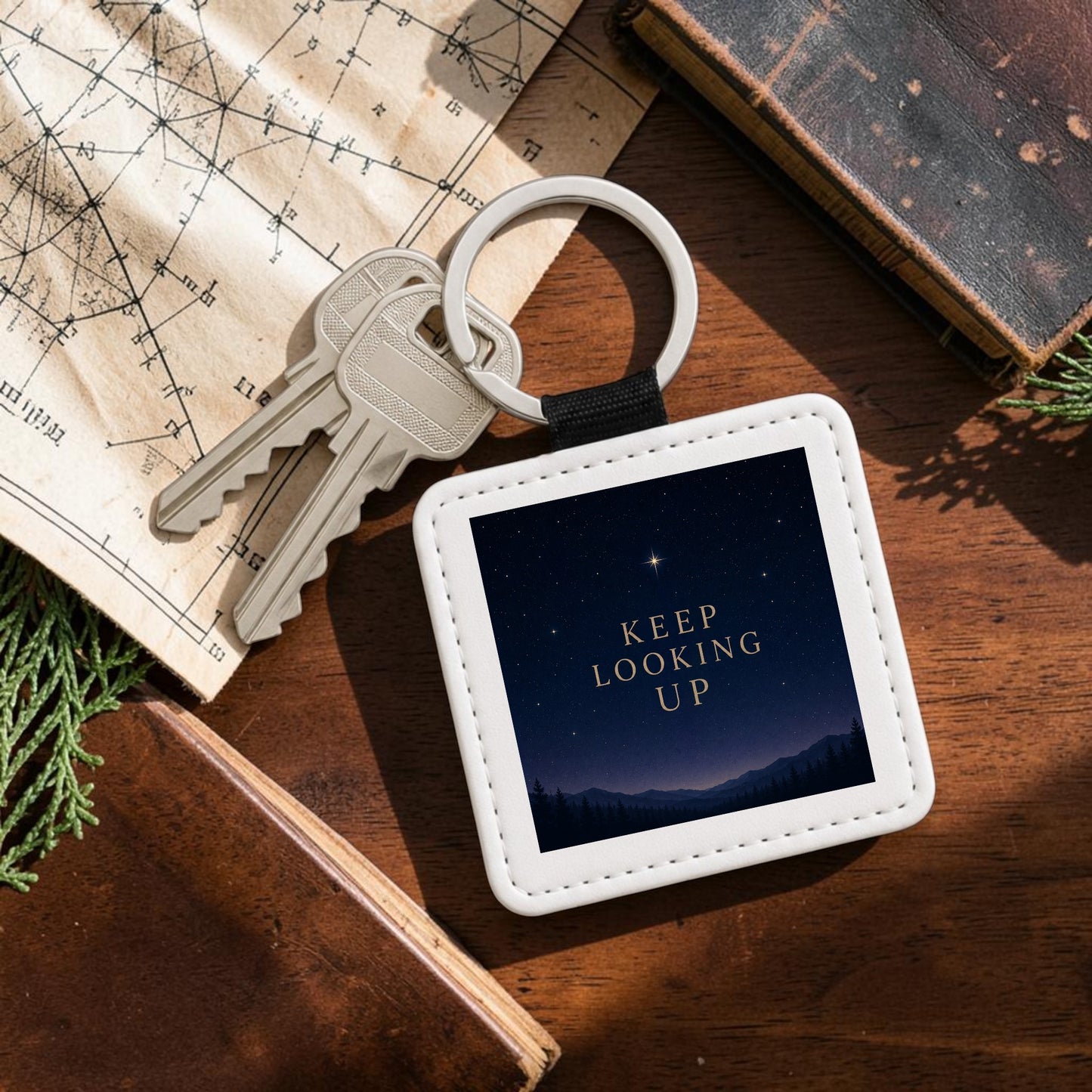 A Keep Looking Up Celestial Keychain by Mirabilia Boutique, featuring an image of a night sky with a single bright star and the words “KEEP LOOKING UP,” rests on a table next to a star chart, holding a set of keys.