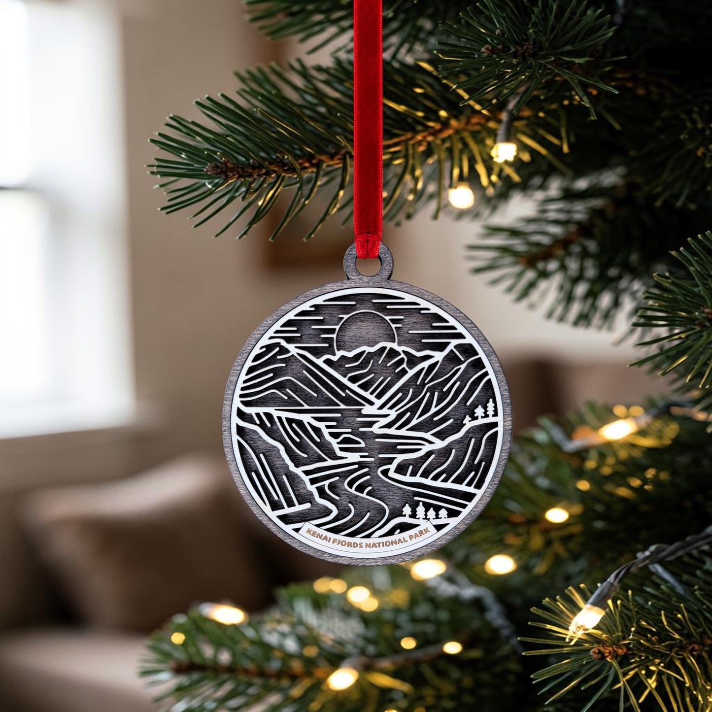 The Kenai Fjords National Park Line Art Christmas Ornament by Mirabilia Boutique is a silver, oval ornament adorned with elegant mountain and river line art. Hanging from a red ribbon on a Christmas tree with white lights, it brings the captivating landscapes of Kenai Fjords National Park into your holiday decor. The softly blurred background reveals part of a window and couch, highlighting this unique and inspiring ornament.