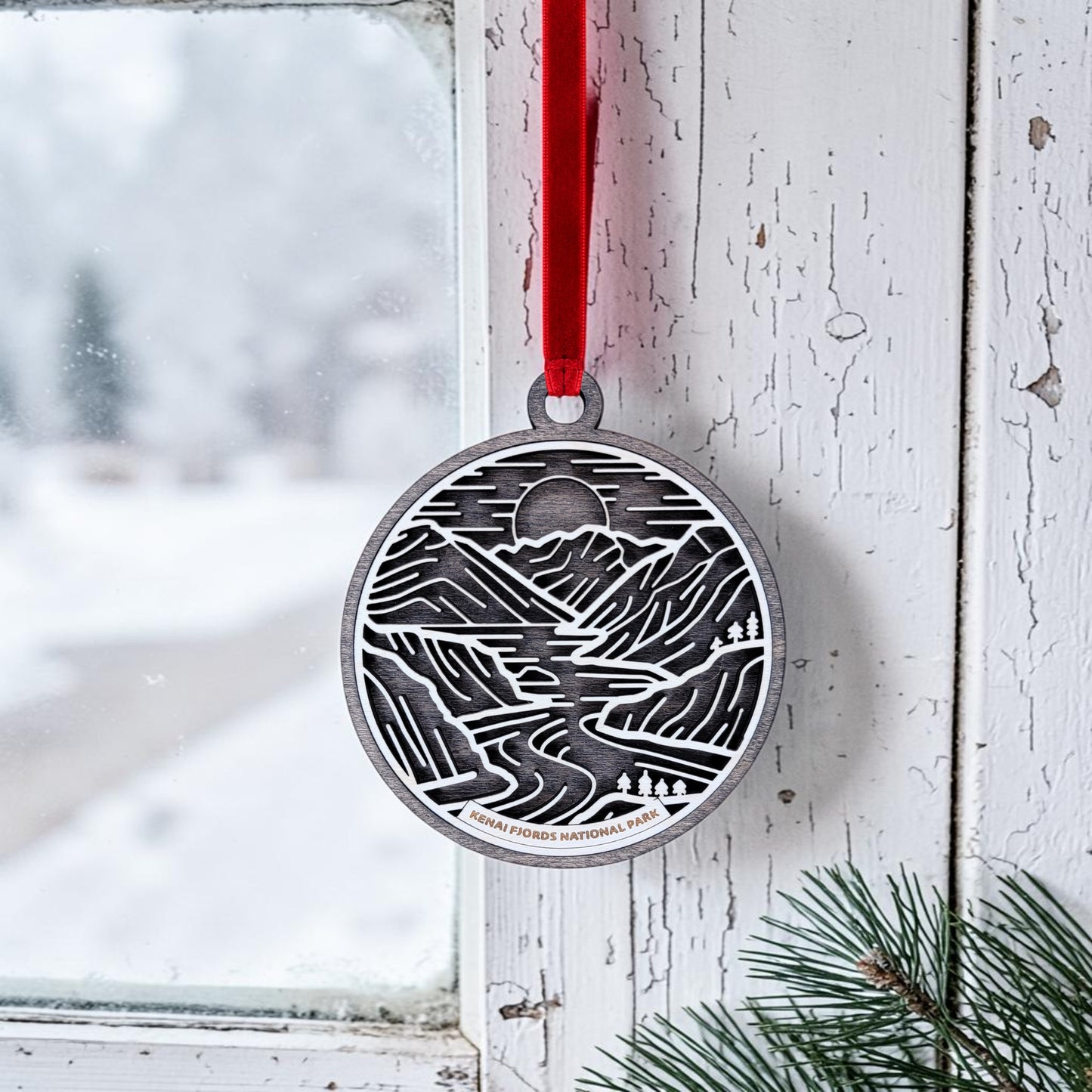The Kenai Fjords National Park Line Art Christmas Ornament by Mirabilia Boutique, featuring a mountain and river design, hangs from a red ribbon on a white window frame. Snow is visible outside, while pine branches rest in the corner.