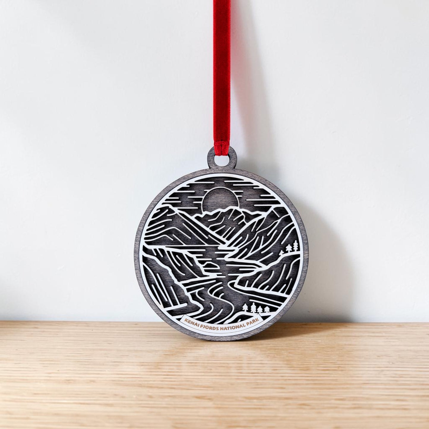 The Kenai Fjords National Park Line Art Christmas Ornament by Mirabilia Boutique, a round metal decoration adorned with elegant engravings of mountains, a river, trees, and a sun inspired by the park's landscapes, hangs from a red ribbon against a light wall and is displayed on a wooden surface.