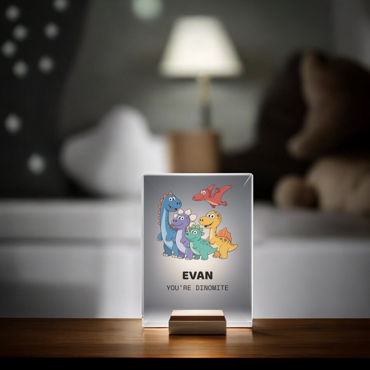 The Mirabilia Boutique Personalized Dinosaur Night Light for Kids sits on a table, featuring cartoon dinosaurs and "EVAN YOU'RE DINOMITE," with a cozy kids' bedroom backdrop of a lamp, couch, and star-decorated wall.
