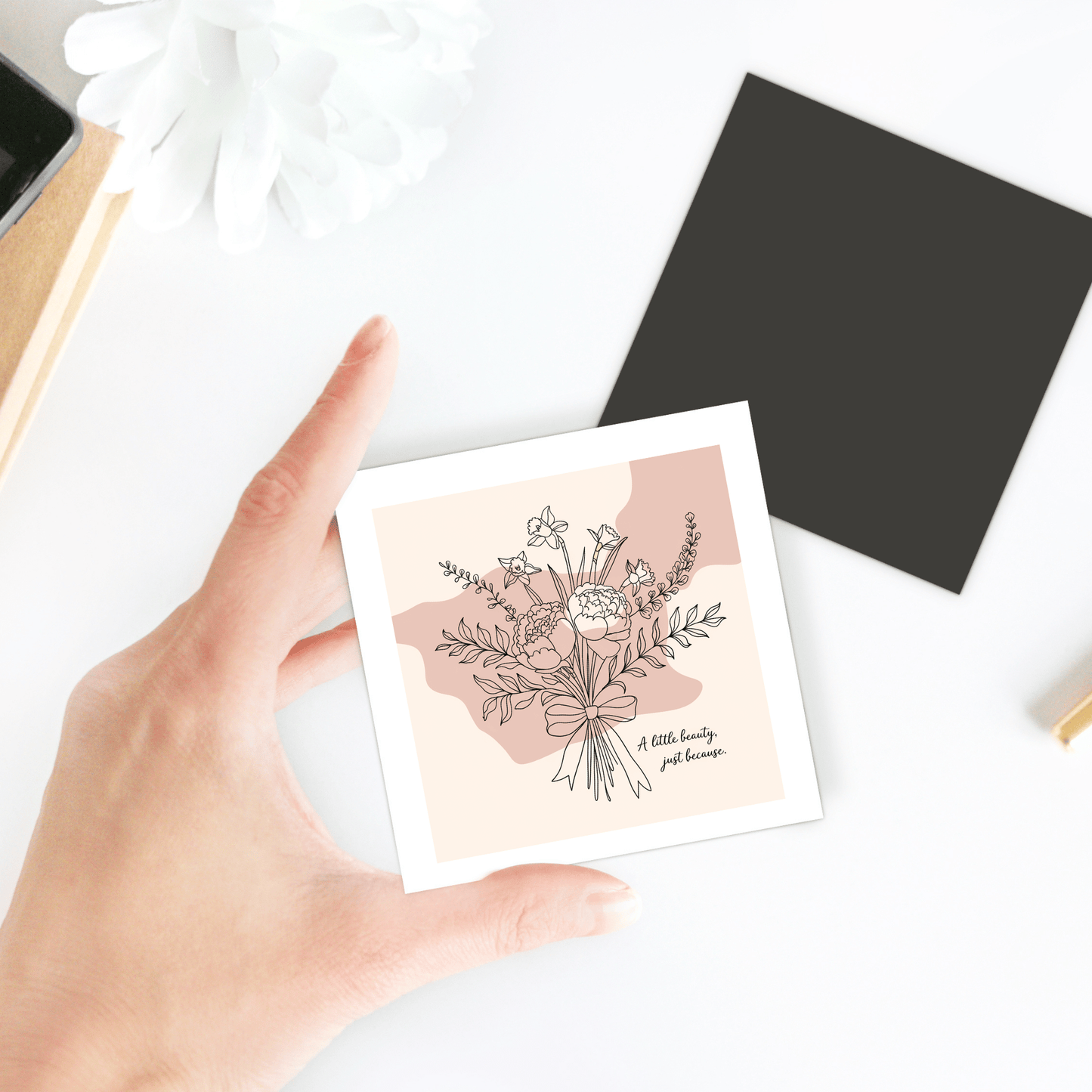 A hand holds the "A Little Beauty Magnet – Just Because Floral Gift" by Mirabilia Boutique, alongside a black square sheet, white flower, and desk items on a white surface.