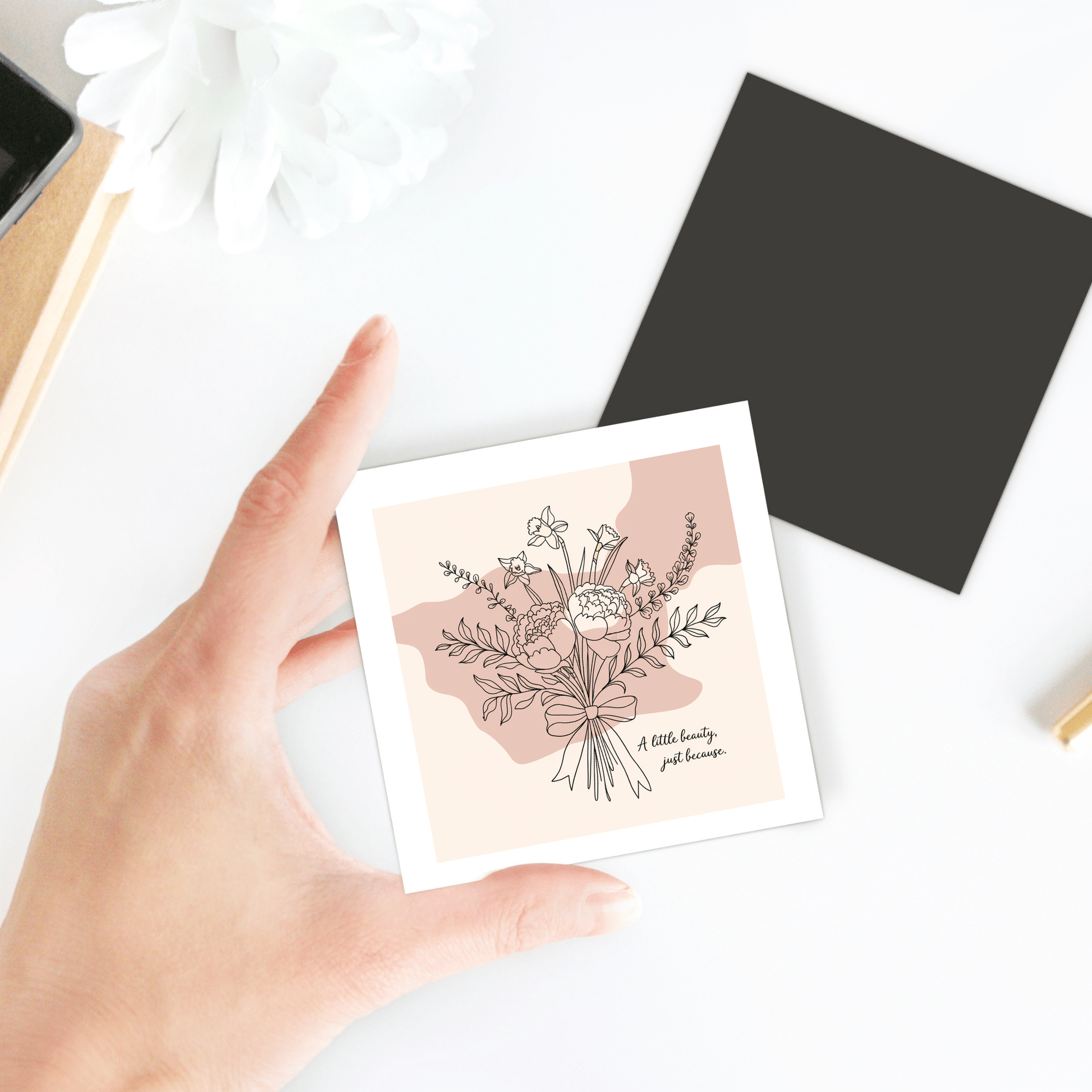 A hand holds the "A Little Beauty Magnet – Just Because Floral Gift" by Mirabilia Boutique, alongside a black square sheet, white flower, and desk items on a white surface.