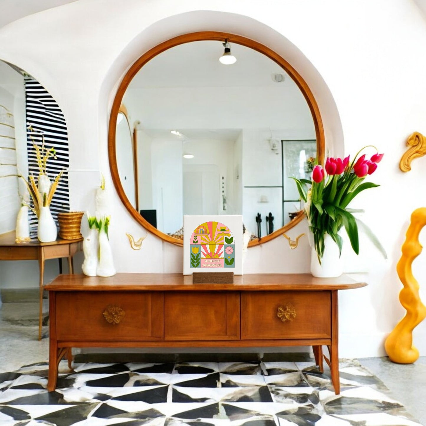 A wooden console table sits beneath a large round mirror, holding a vase of pink tulips, a decorative box, and the Mirabilia Boutique "Radiate Goodness" mini canvas sign. White walls and a black-and-white geometric floor complete the modern look.
