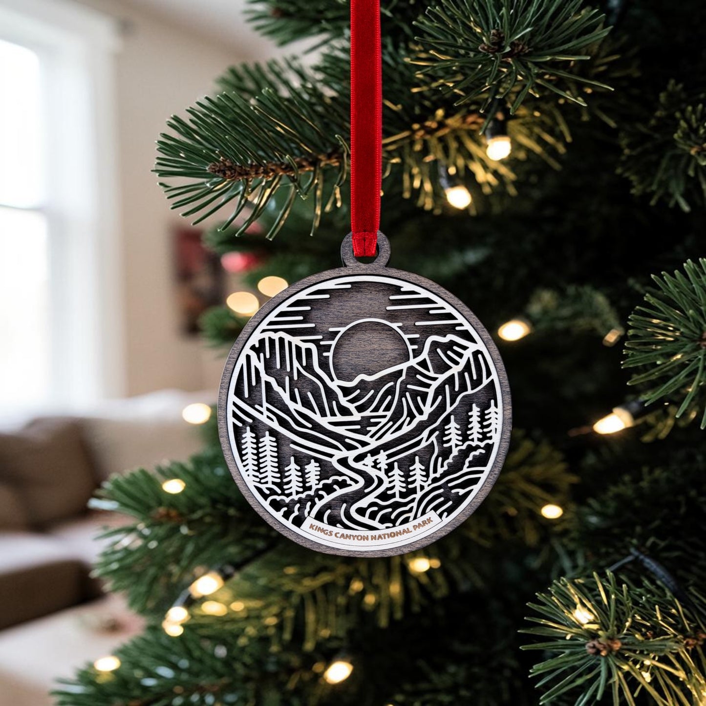 The Kings Canyon National Park Line Art Christmas Ornament by Mirabilia Boutique—a round wooden ornament depicting a mountain and forest scene inspired by the giant sequoias—hangs from a red ribbon on a Christmas tree adorned with white lights, while in the background, a cozy room with a window and couch is visible.