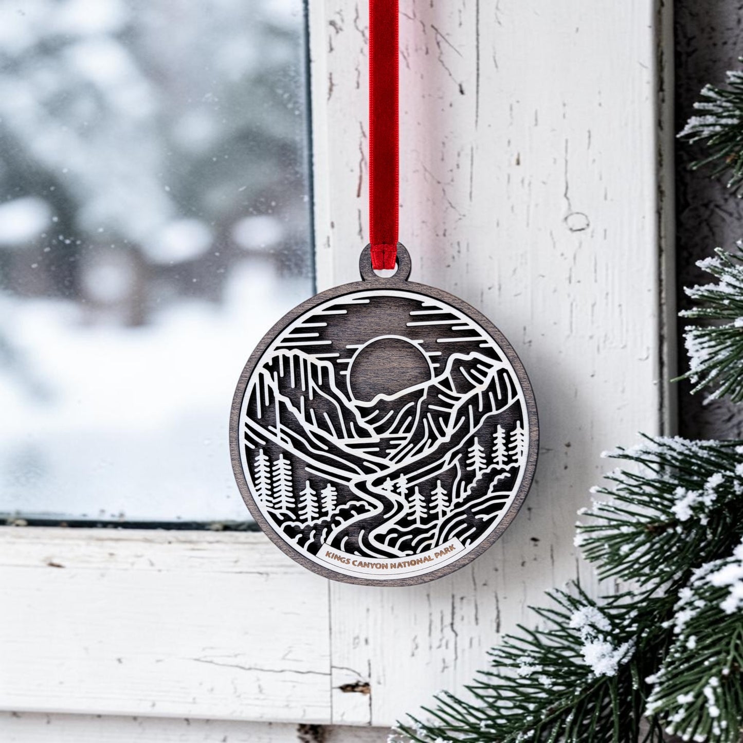 The Kings Canyon National Park Line Art Christmas Ornament by Mirabilia Boutique—a round wooden ornament depicting a mountain and forest scene inspired by the giant sequoias of Kings Canyon—hangs from a red ribbon on a snowy window, surrounded by green pine branches and fresh snow.