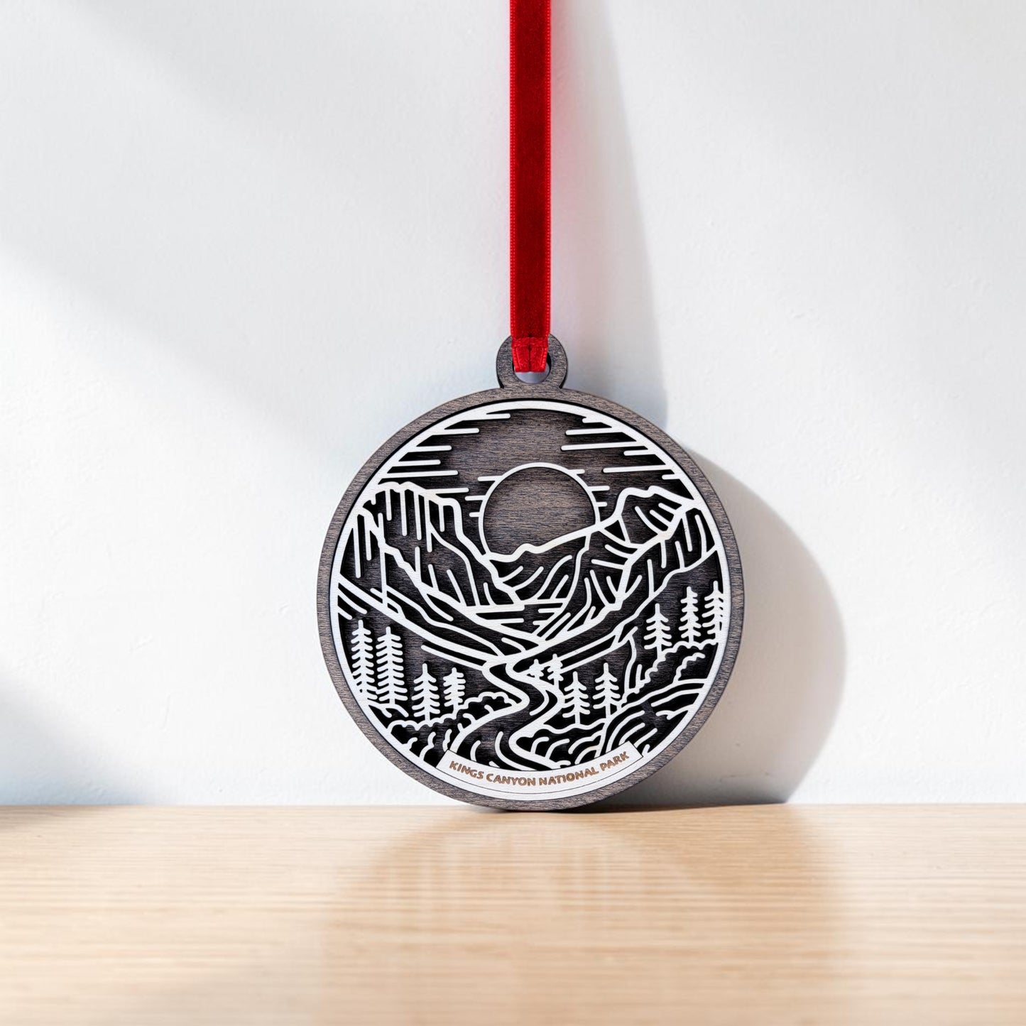 The Kings Canyon National Park Line Art Christmas Ornament by Mirabilia Boutique features a round wooden design, laser-cut with mountains, sequoia-inspired trees, and a flowing river; it hangs from a red ribbon against a white wall atop a light wooden surface, beautifully capturing the natural essence of Kings Canyon National Park.