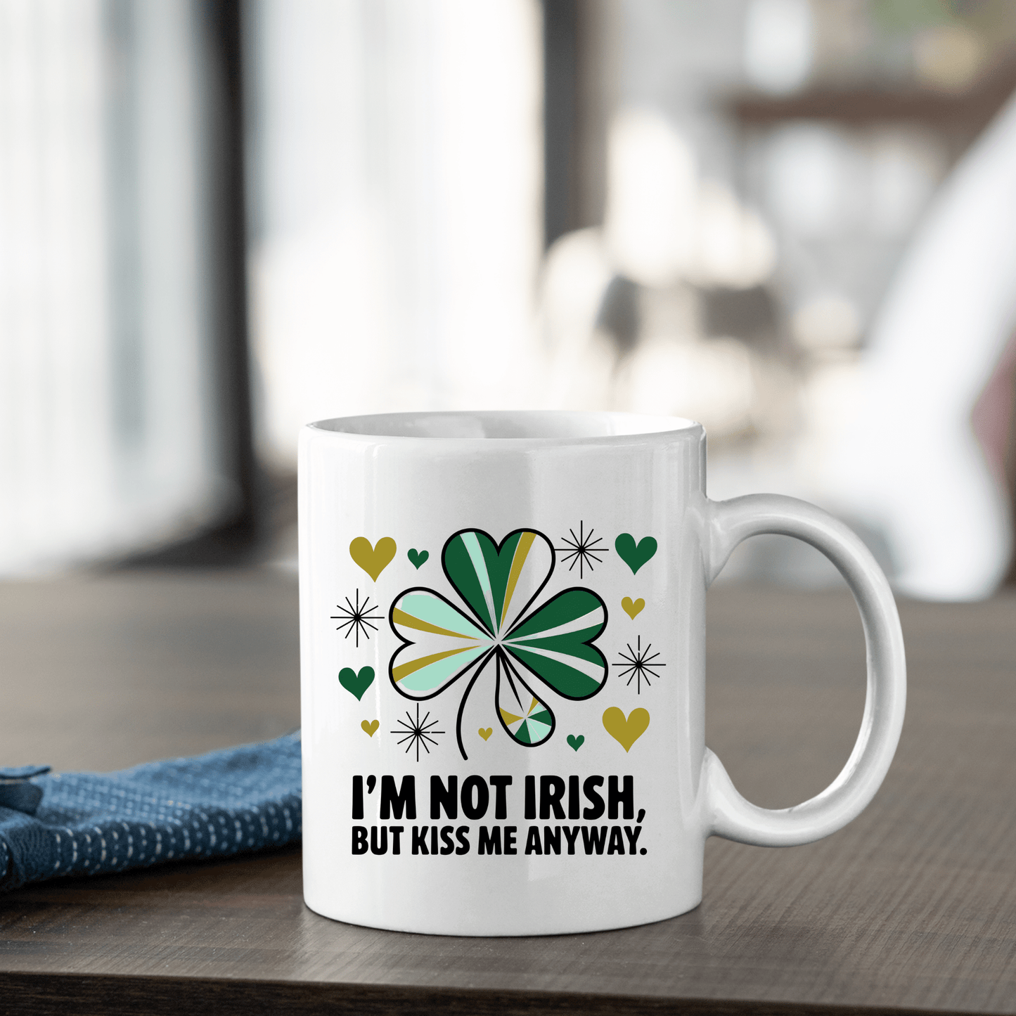 The Kiss Me Anyway Mug – St. Patrick’s Day Charm by Mirabilia Boutique sits on a wooden table, featuring a green shamrock, yellow and green hearts, and “I’M NOT IRISH, BUT KISS ME ANYWAY.” A folded blue cloth rests nearby.