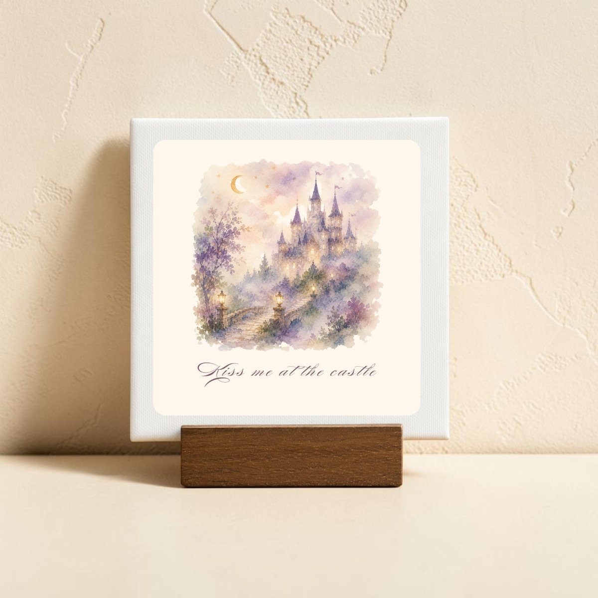 The Kiss Me at the Castle Mini Canvas Sign by Mirabilia Boutique features a dreamy watercolor castle scene with a crescent moon and delicate flowers, evoking fairytale romance. Displayed on a wooden stand, this enchanting piece of castle home decor showcases the phrase "Kiss me at the castle" beneath the artwork. Resting on a light surface against a textured beige wall, it makes an ideal decorative accent for any space.