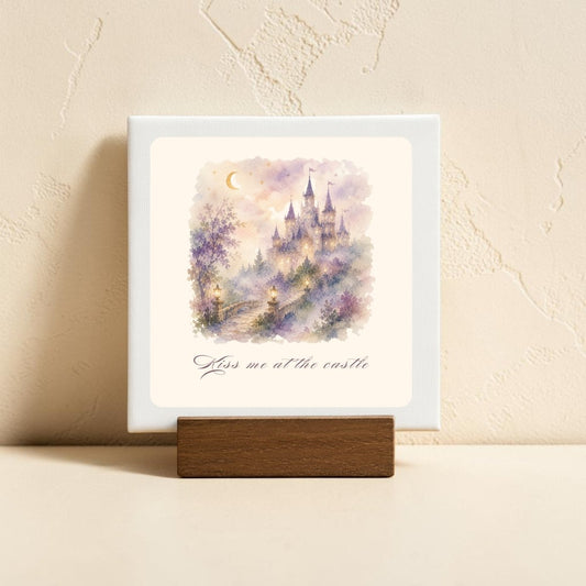 The Kiss Me at the Castle Mini Canvas Sign by Mirabilia Boutique features a dreamy watercolor castle scene with a crescent moon and delicate flowers, evoking fairytale romance. Displayed on a wooden stand, this enchanting piece of castle home decor showcases the phrase "Kiss me at the castle" beneath the artwork. Resting on a light surface against a textured beige wall, it makes an ideal decorative accent for any space.