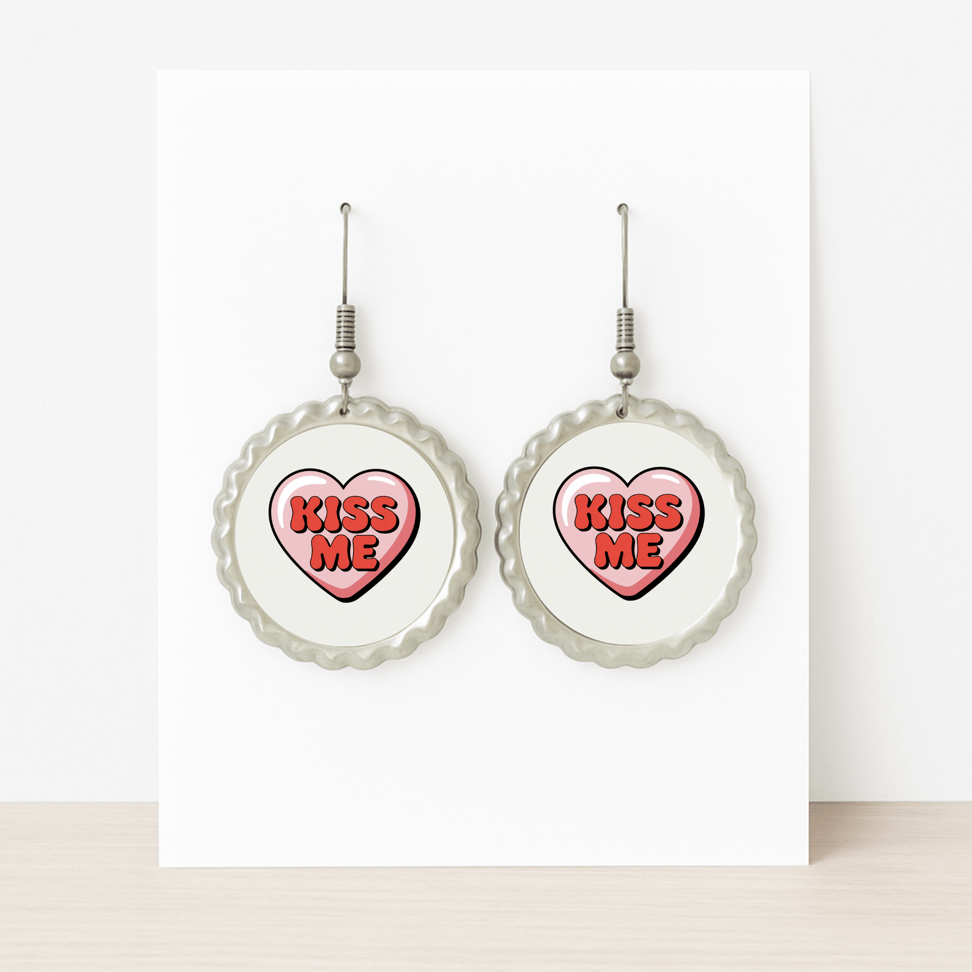 The Kiss Me Heart Earrings by Mirabilia Boutique feature round studs with heart-shaped centers reading "KISS ME" in bold red—an ideal Valentine’s Day gift, displayed on a white card atop a light wood surface.