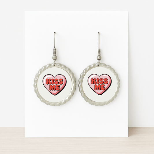 The Kiss Me Heart Earrings by Mirabilia Boutique feature round studs with heart-shaped centers reading "KISS ME" in bold red—an ideal Valentine’s Day gift, displayed on a white card atop a light wood surface.