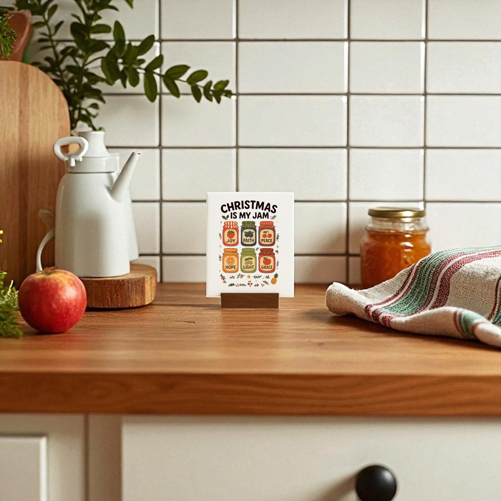 A kitchen scene with a wooden counter featuring an apple, a white teapot, a jar of jam, a striped towel, and the Mirabilia Boutique "Christmas Is My Jam" festive holiday table decor sign.