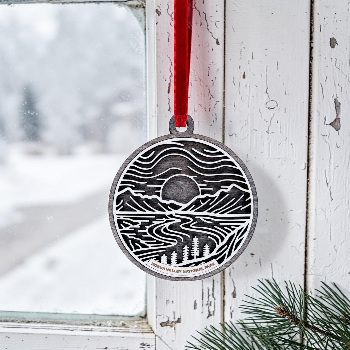 A round metal ornament from Mirabilia Boutique, the Kobuk Valley National Park Line Art Christmas Ornament, hangs in a window by a red ribbon. Snow drifts gently outside while pine branches peek from the lower right corner, creating a wintry scene that beautifully captures the landscape charm of Alaska’s Kobuk Valley.