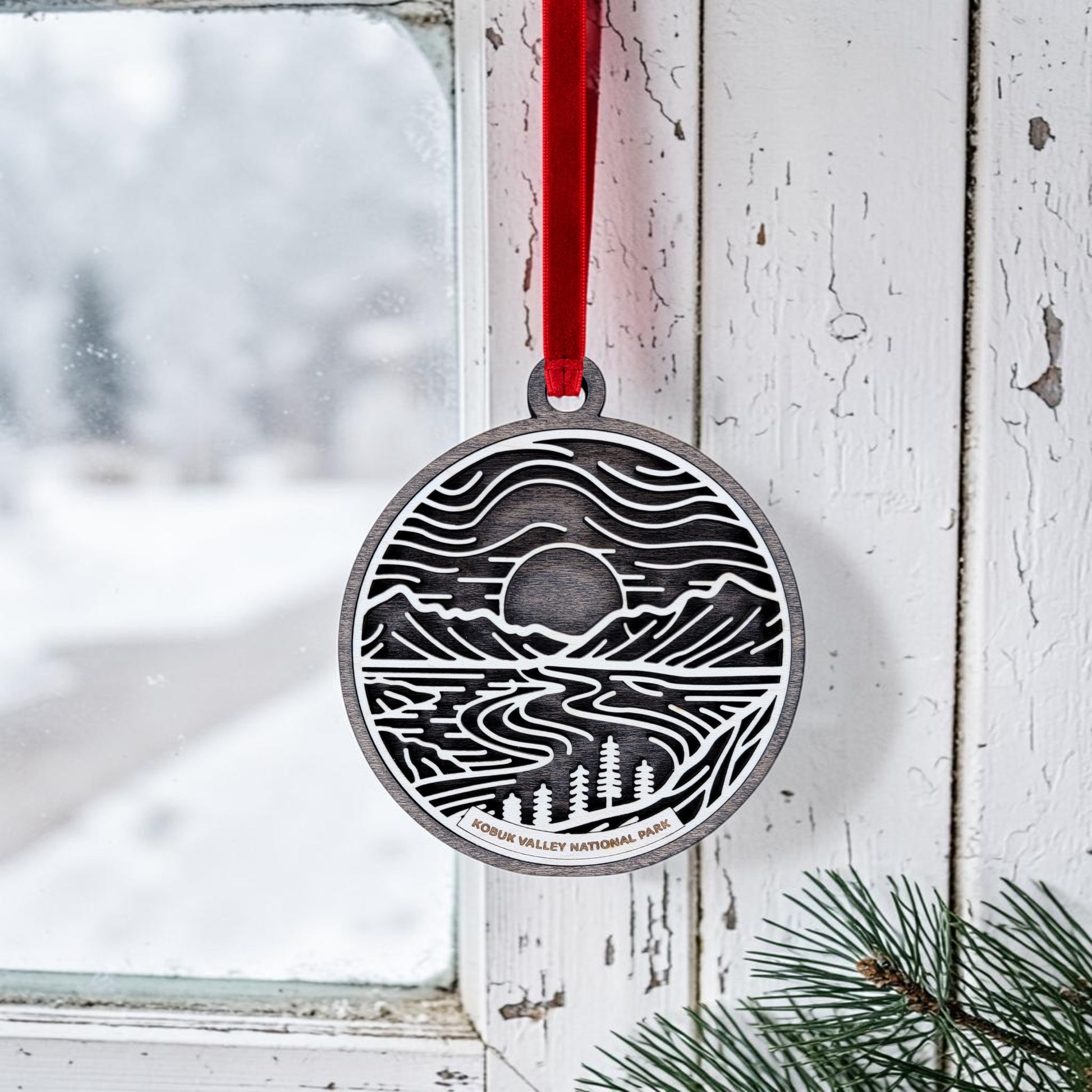 A round metal ornament from Mirabilia Boutique, the Kobuk Valley National Park Line Art Christmas Ornament, hangs in a window by a red ribbon. Snow drifts gently outside while pine branches peek from the lower right corner, creating a wintry scene that beautifully captures the landscape charm of Alaska’s Kobuk Valley.