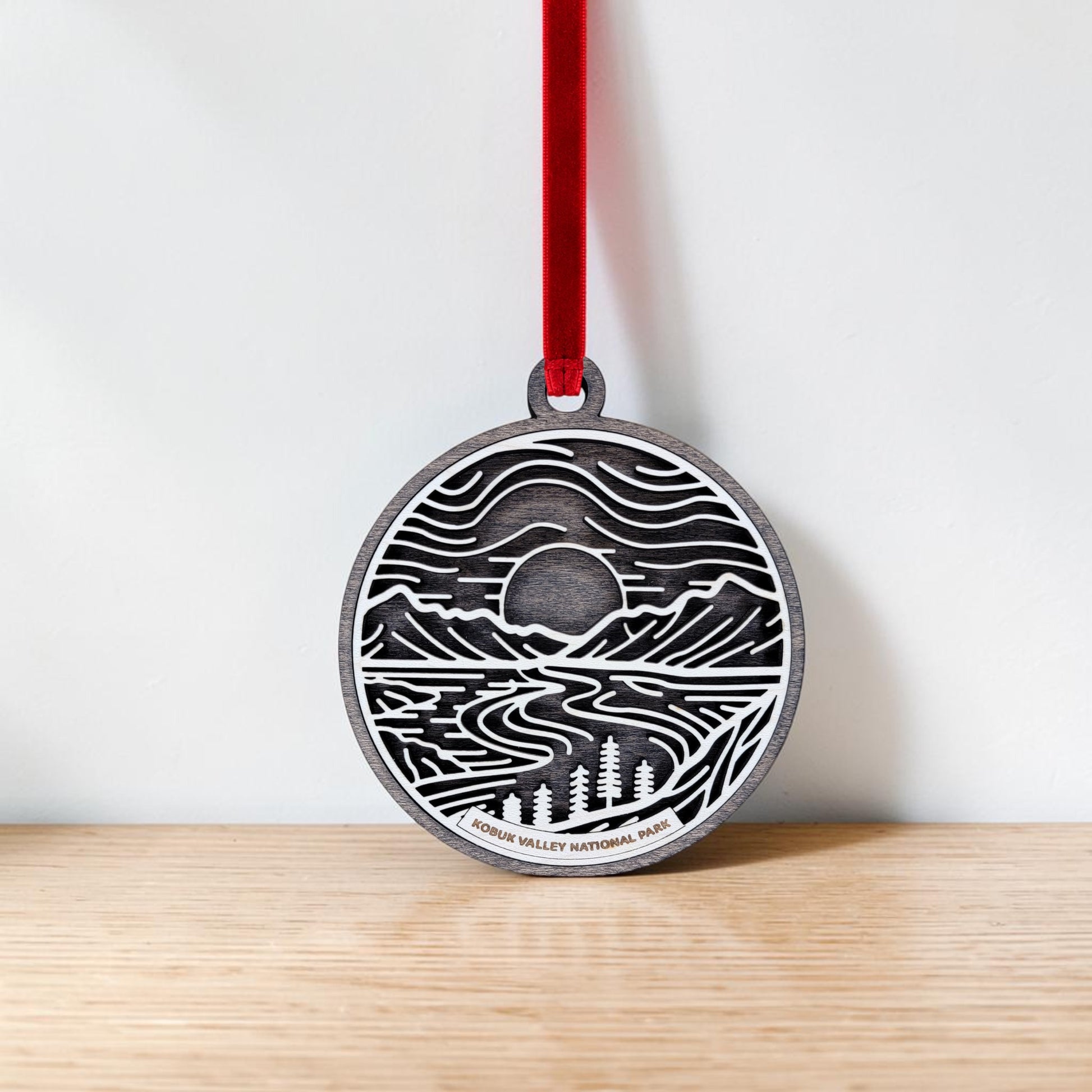 A round wooden ornament from Mirabilia Boutique hangs on a red ribbon, featuring line art of mountains, trees, a winding river, and a setting sun reminiscent of Alaska. Labeled “Kobuk Valley National Park,” this handcrafted Christmas ornament rests against a light wood background.