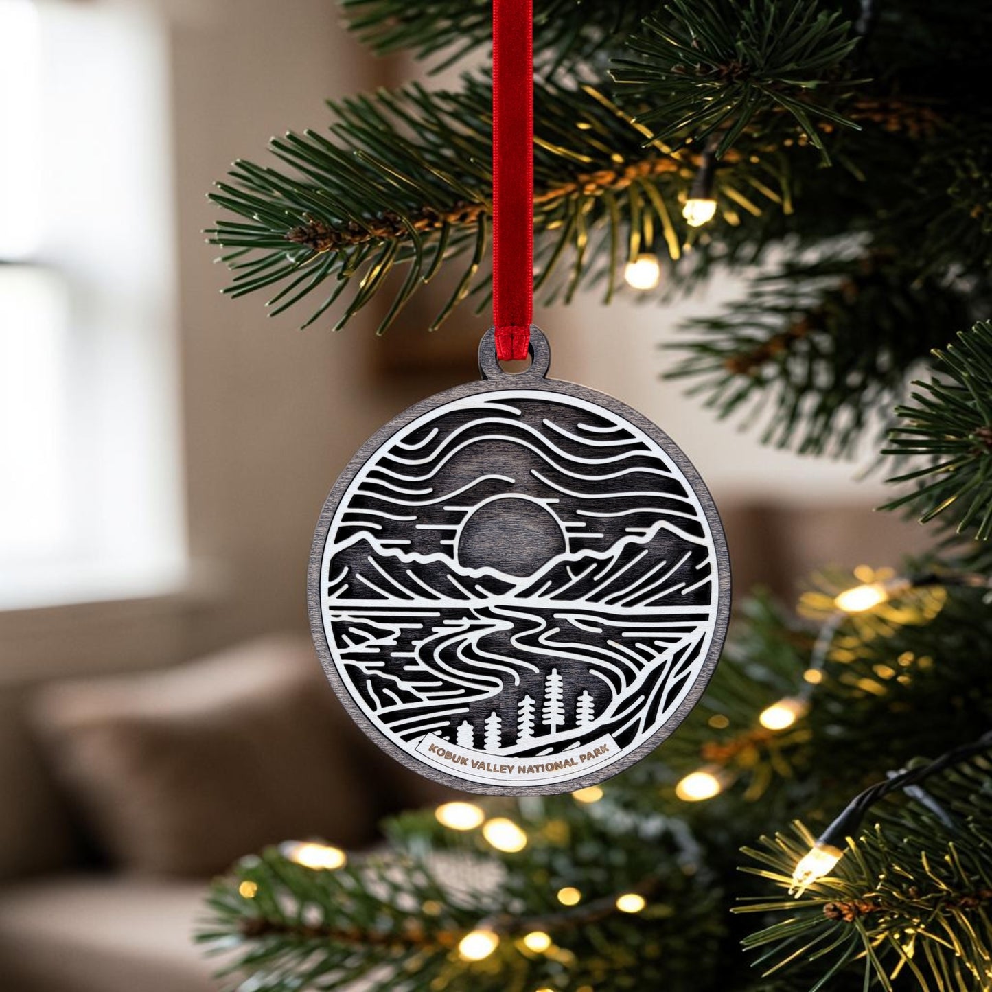 A round metal ornament from Mirabilia Boutique, featuring a Kobuk Valley National Park line art design, hangs by a red ribbon on a Christmas tree adorned with small yellow lights. In the background, a softly blurred living room creates an inviting setting to showcase this special Alaska keepsake.