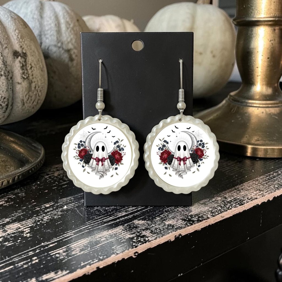 Two round Crescent Moon Lace Ghost Scalloped Earrings from Mirabilia Boutique, featuring ghost faces, red roses, and black leaf motifs, are displayed on a black card. Styled with vintage-inspired scalloped crescent moons for an enchanting touch, this jewelry piece exudes mystery and elegance. In the background, white pumpkins and a brass candlestick rest on a dark wooden surface, enhancing the captivating ambiance.
