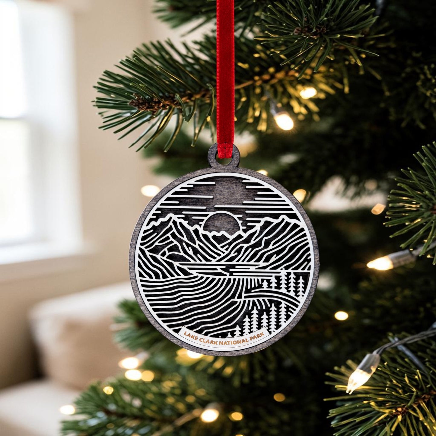 The Lake Clark National Park Line Art Christmas Ornament by Mirabilia Boutique, featuring a detailed mountain landscape and "Lake Clark National Park" text, hangs beautifully on a Christmas tree with white lights—making it the ideal Alaska wilderness keepsake.