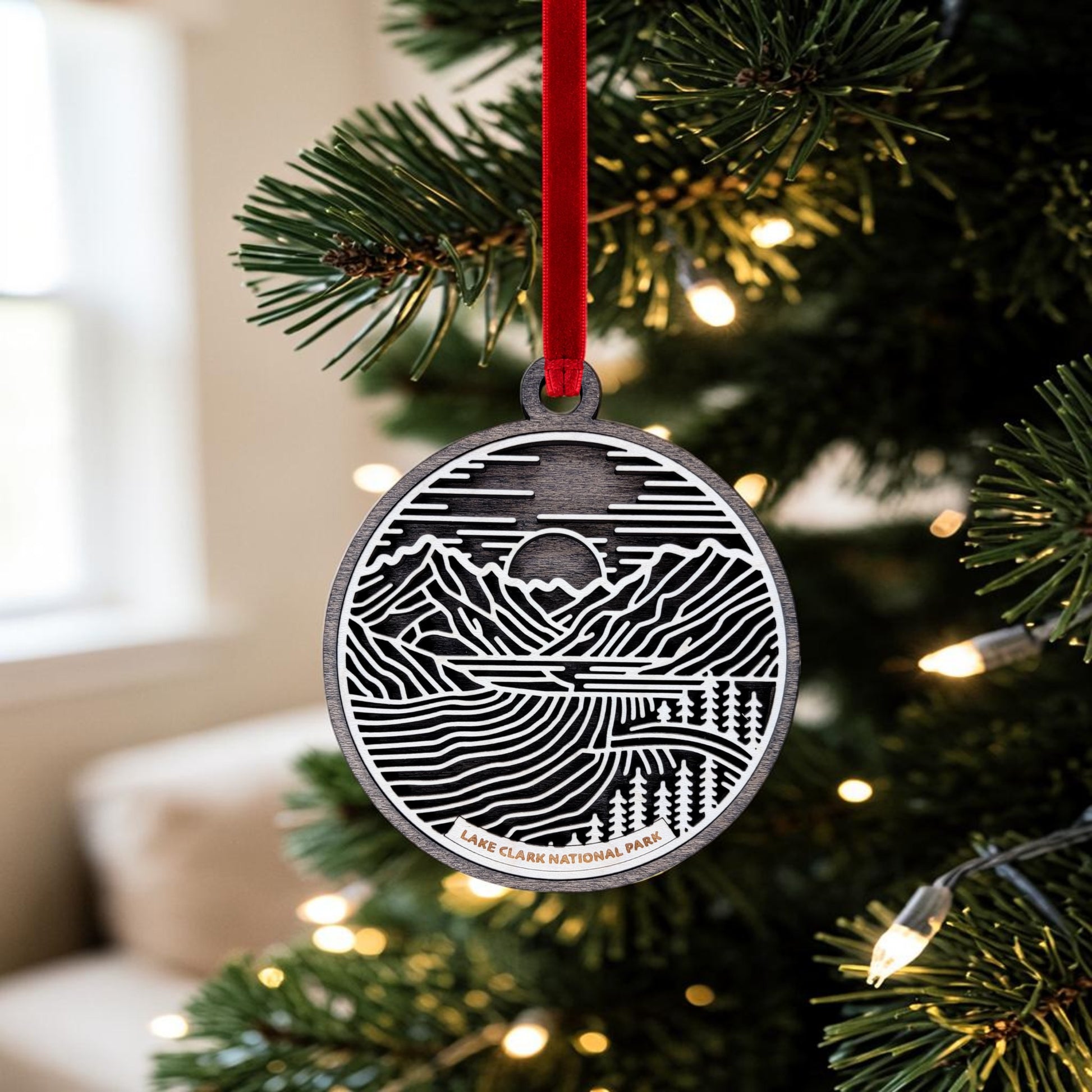 The Lake Clark National Park Line Art Christmas Ornament by Mirabilia Boutique, featuring a detailed mountain landscape and "Lake Clark National Park" text, hangs beautifully on a Christmas tree with white lights—making it the ideal Alaska wilderness keepsake.