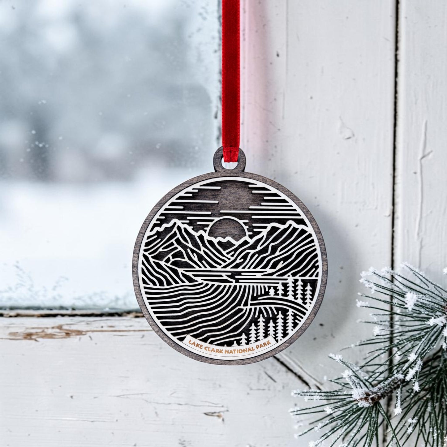 A handcrafted Lake Clark National Park Line Art Christmas Ornament by Mirabilia Boutique, featuring a detailed mountain and lake scene, hangs by a red ribbon on a frosty window beside pine branches. This Alaska wilderness keepsake beautifully reflects the snowy landscape outside.