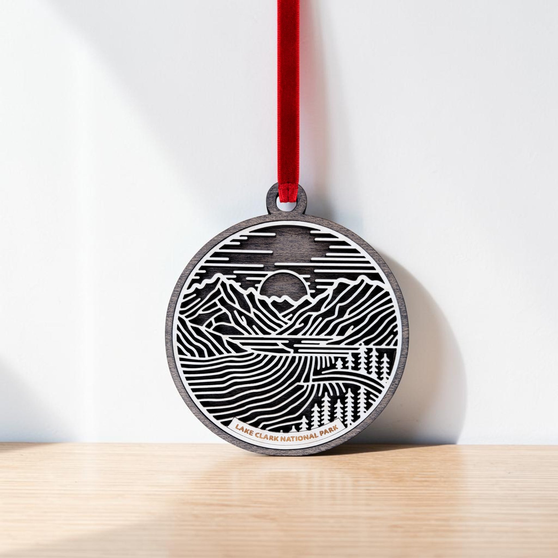 Handcrafted by Mirabilia Boutique, the Lake Clark National Park Line Art Christmas Ornament features a mountain and lake landscape design and hangs by a red ribbon against a white wall. "Lake Clark National Park" is engraved at the bottom, making it an ideal keepsake for nature enthusiasts.