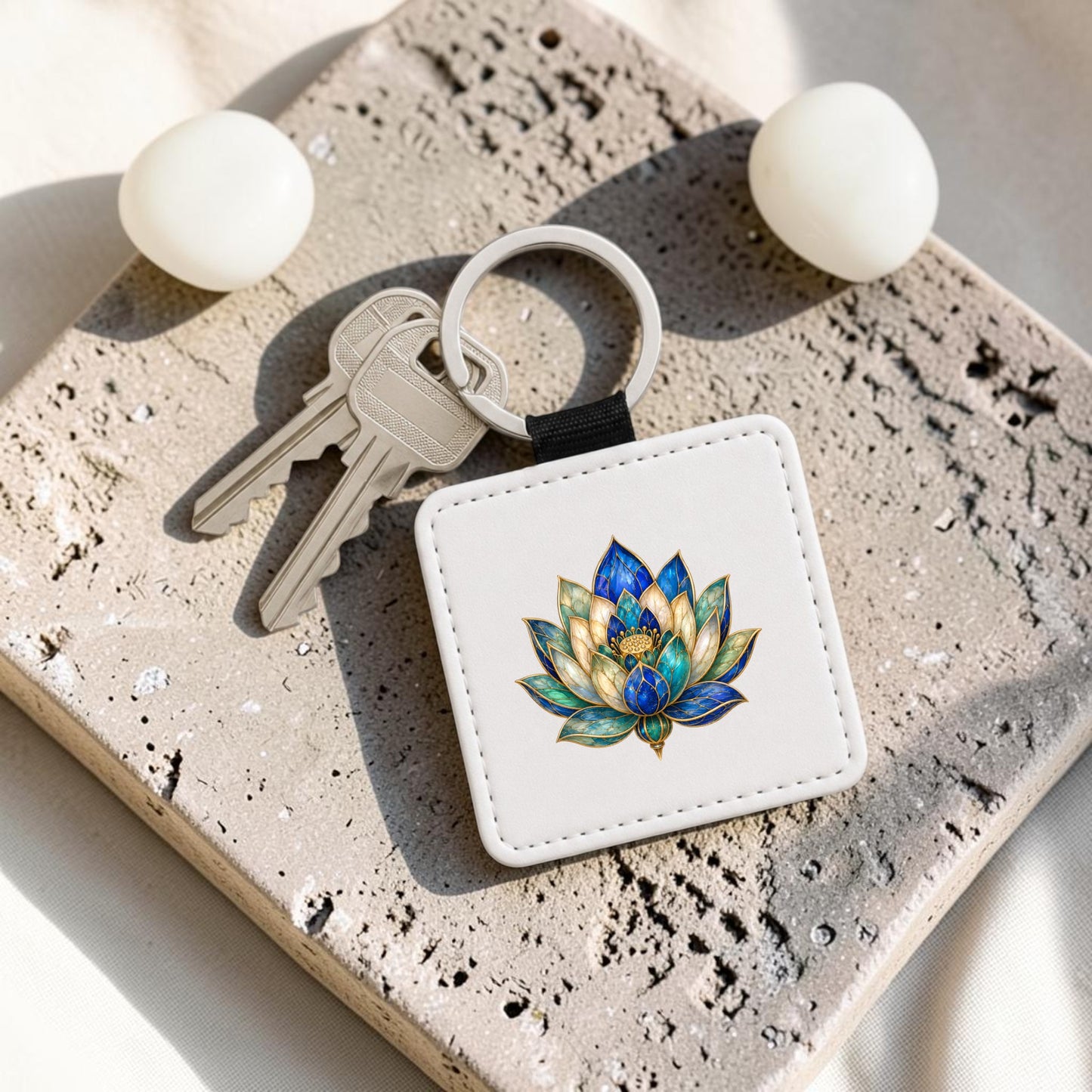 Two silver keys are attached to the Lapis Bloom Keychain from Mirabilia Boutique, a square white accessory featuring vibrant Egyptian lotus art with accents of lapis blue. The keychain rests on a textured stone surface beside two round white objects.