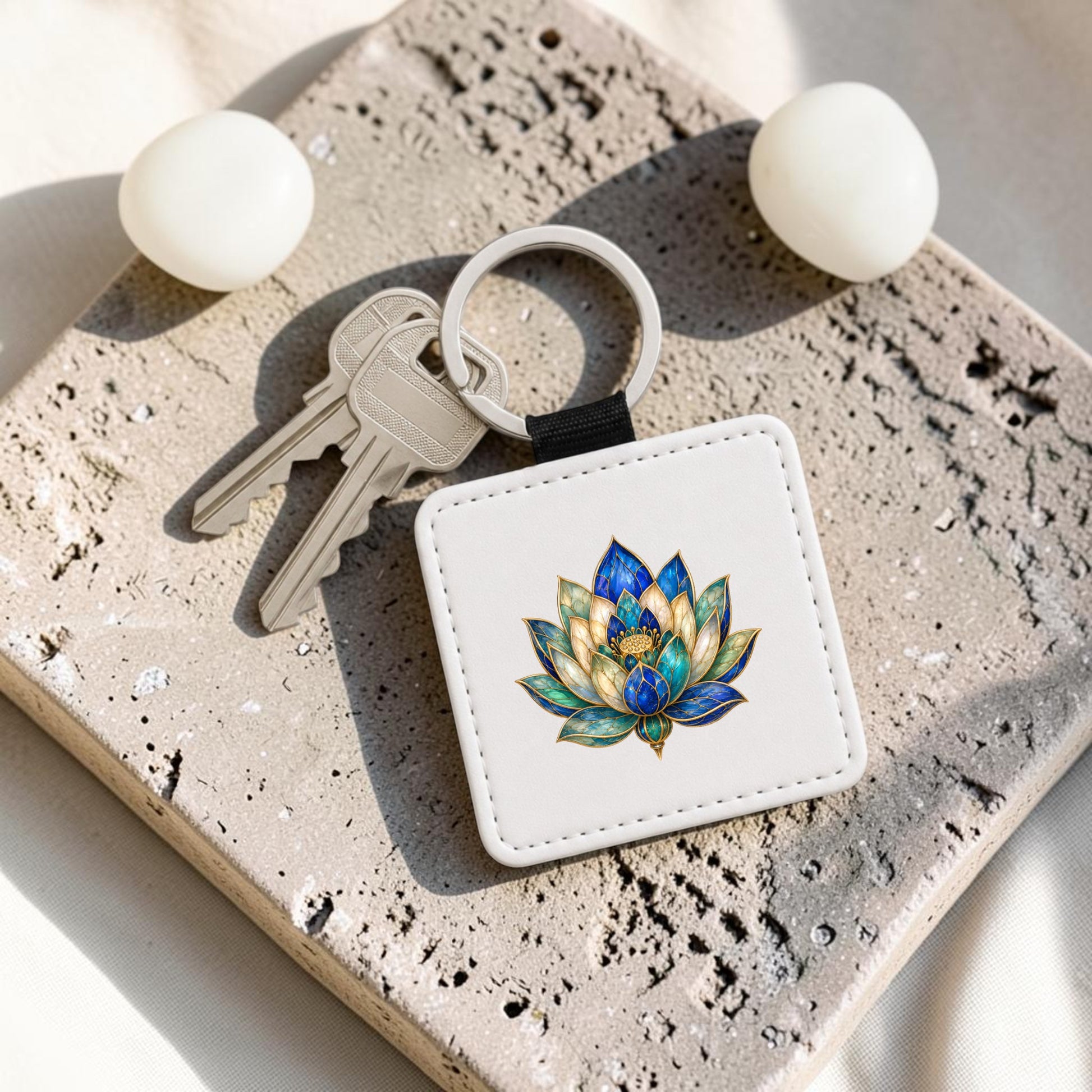 Two silver keys are attached to the Lapis Bloom Keychain from Mirabilia Boutique, a square white accessory featuring vibrant Egyptian lotus art with accents of lapis blue. The keychain rests on a textured stone surface beside two round white objects.