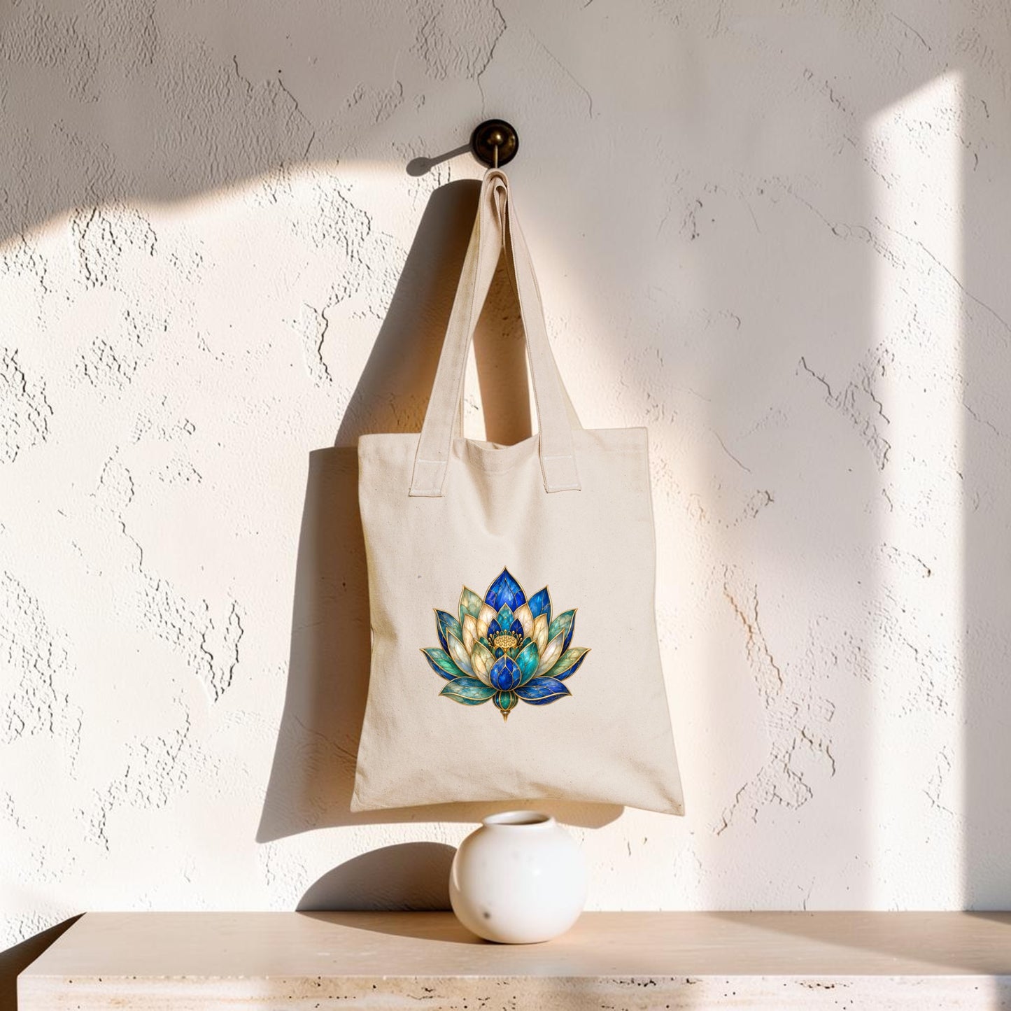 A Lapis Bloom Tote Bag | Egyptian Lotus Art from Mirabilia Boutique, featuring a colorful lotus flower design on beige fabric, hangs on a wall hook above a small white vase, with sunlight casting shadows across the textured wall.