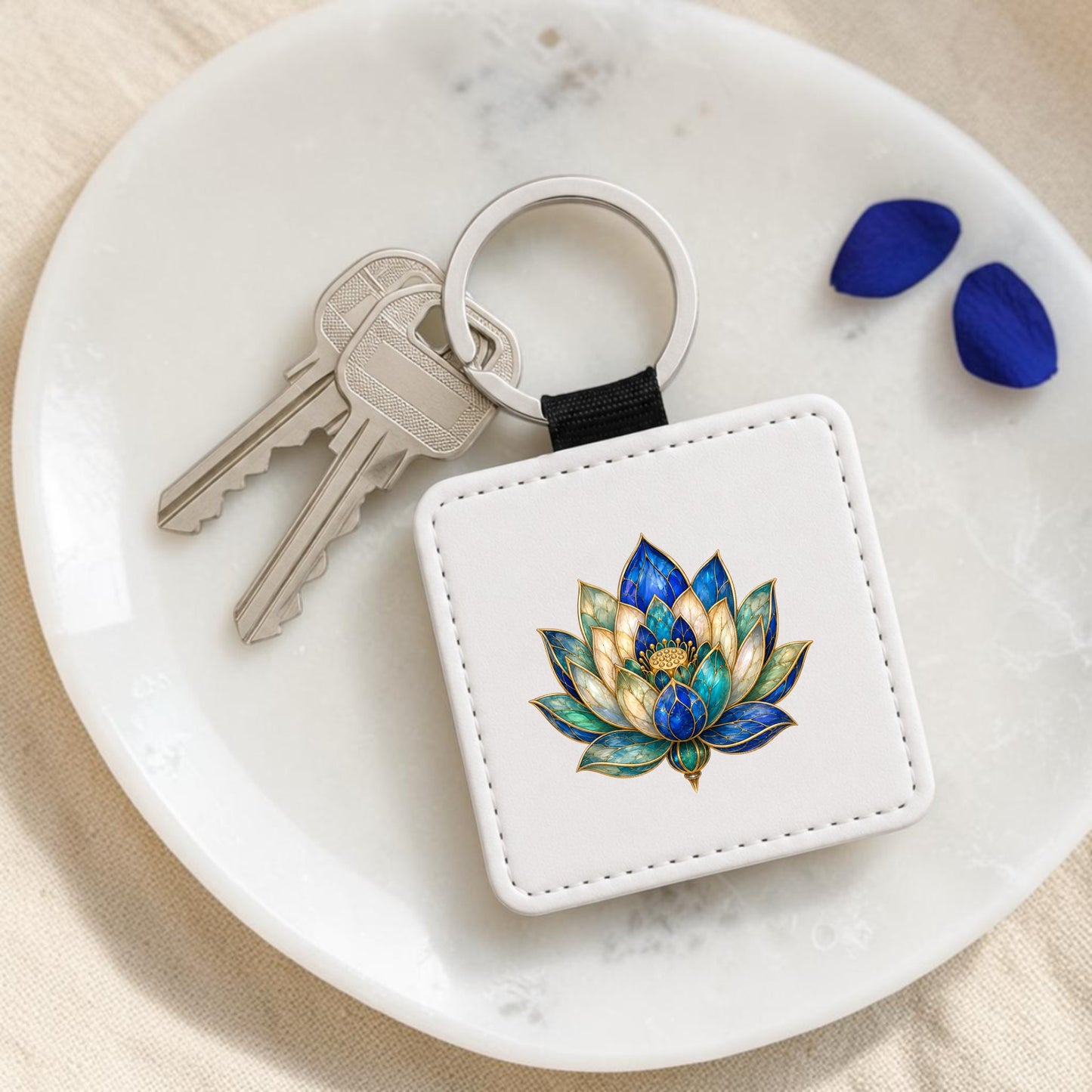 A Lapis Bloom Keychain | Egyptian Lotus Art by Mirabilia Boutique, featuring a colorful lotus design, rests on a white dish beside two keys and two lapis blue petals.
