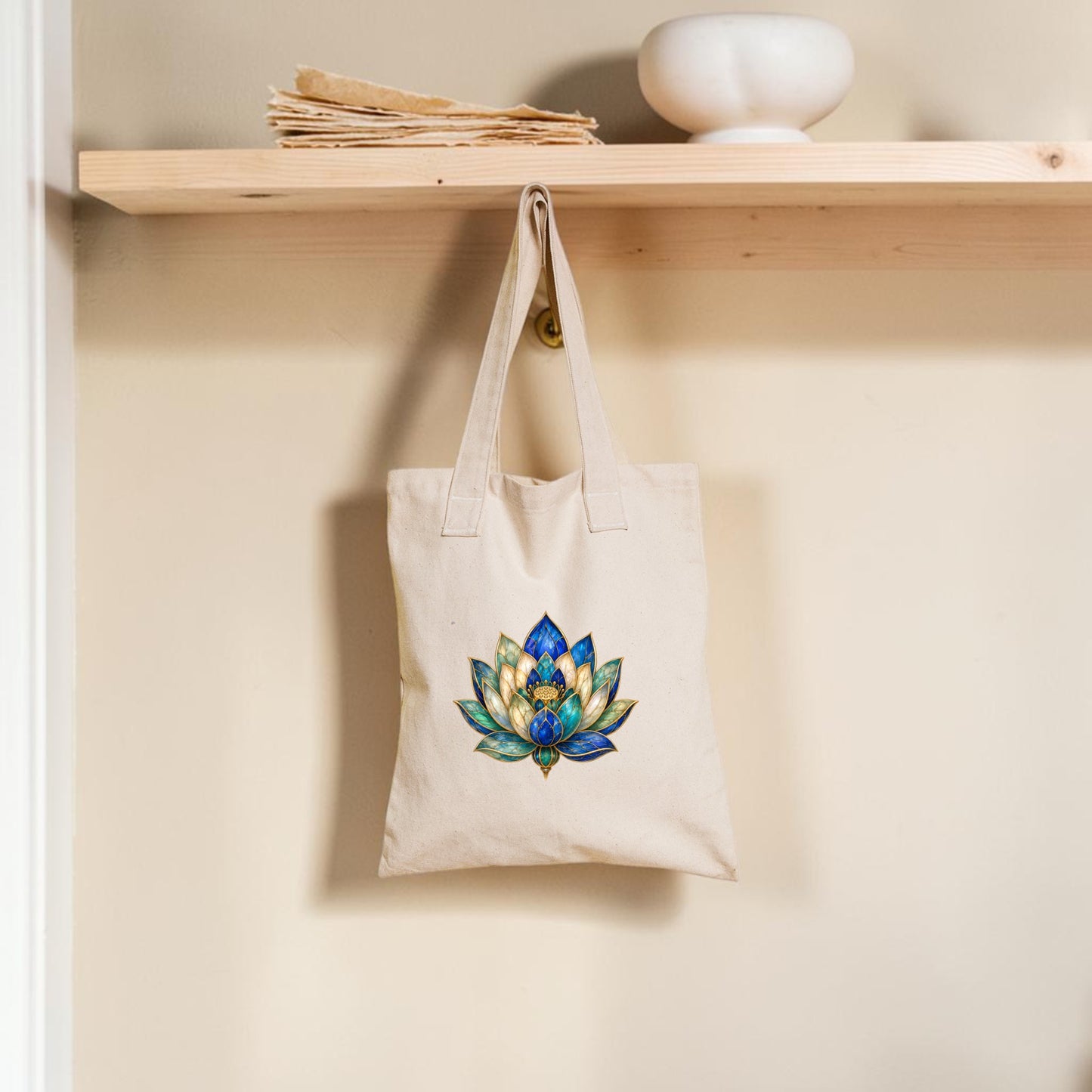 A Lapis Bloom Tote Bag | Egyptian Lotus Art by Mirabilia Boutique, featuring a colorful lotus flower design in beige, hangs from a wooden hook beneath a shelf with papers and a round white bowl against a cream wall.