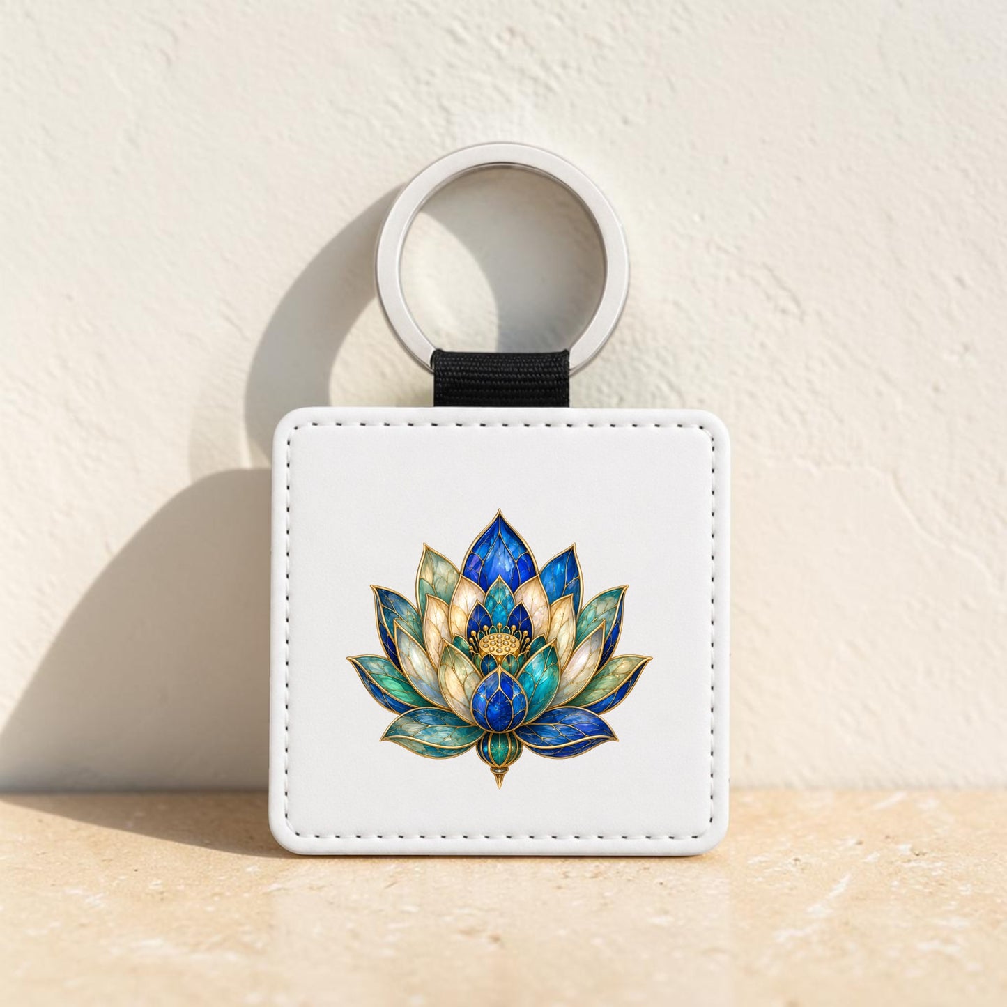 The Lapis Bloom Keychain | Egyptian Lotus Art by Mirabilia Boutique, a white square keychain with a ring adorned with an intricate lapis blue and gold lotus flower design, is displayed upright against a textured light wall on a beige surface.
