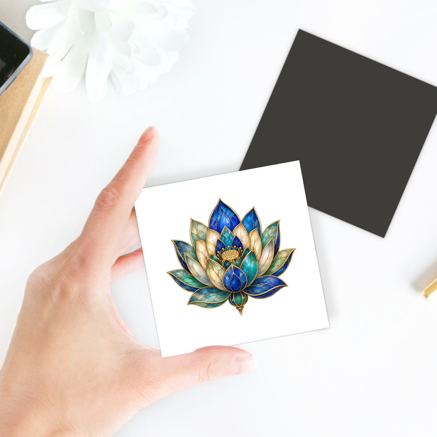 A hand holds the Lapis Bloom Magnet | Egyptian Lotus Art by Mirabilia Boutique, featuring a blue and gold lotus lapis flower illustration on a white card. Also visible on the white surface are a black square card, a flower, and part of a notebook, creating an elegant botanical decor arrangement.