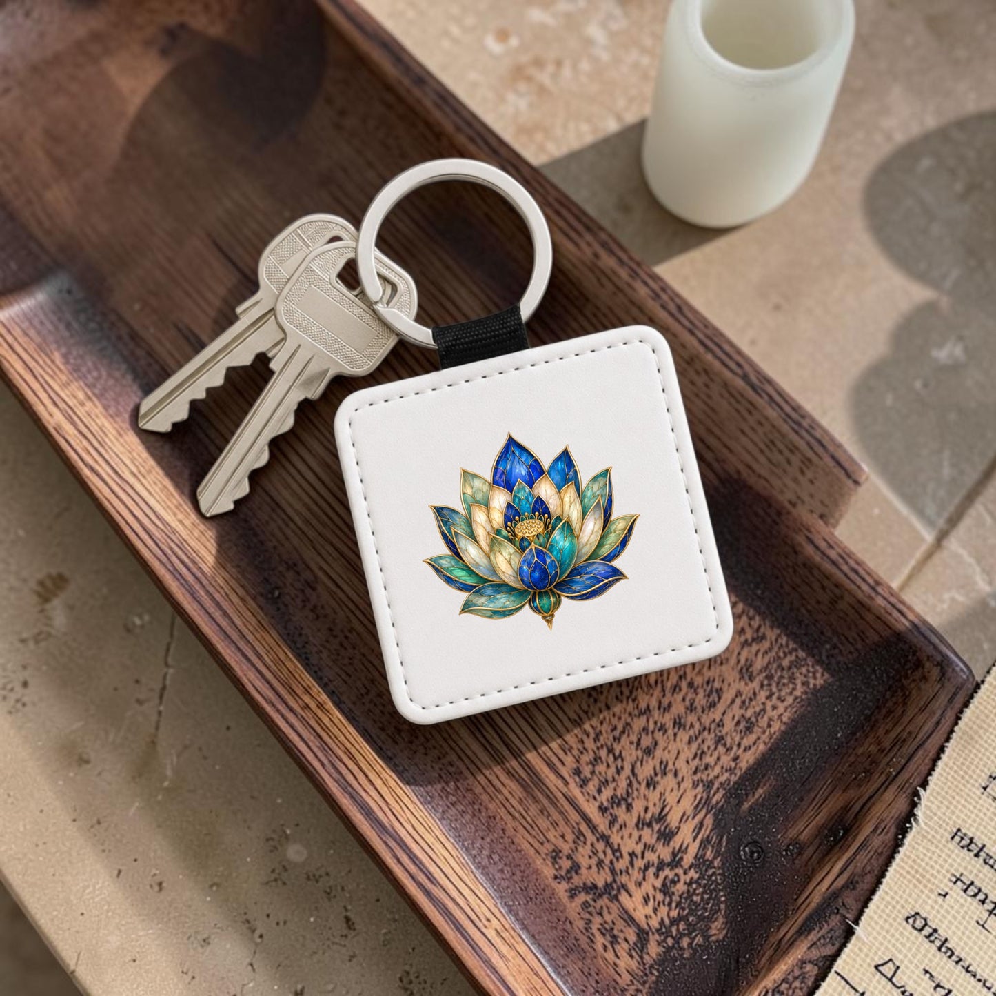 A set of keys with the Lapis Bloom Keychain by Mirabilia Boutique—featuring a colorful Egyptian lotus flower design in shades of lapis blue—rests in a wooden tray beside a small white candle.