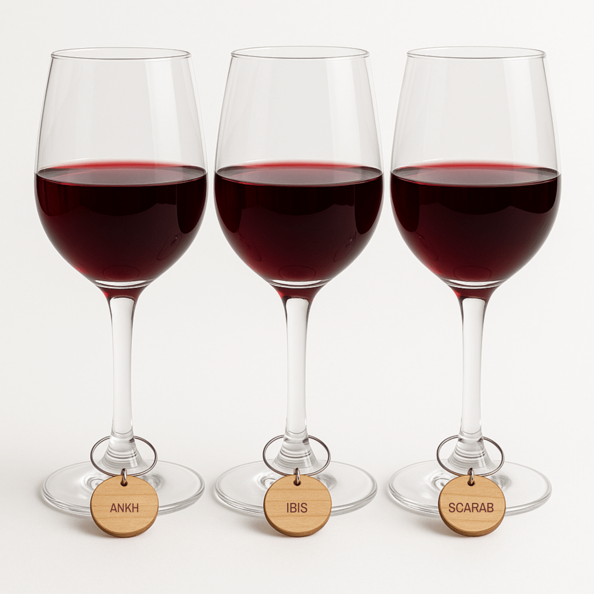 Three wine glasses filled with red wine, each featuring a wooden stem tag labeled “ANKH,” “IBIS,” and “SCARAB” from left to right, display the Ancient Egyptian Wine Charms by Mirabilia Boutique (Lapis & Lotus Collection)—a captivating charm set inspired by Ancient Egyptian motifs—against a plain white background.