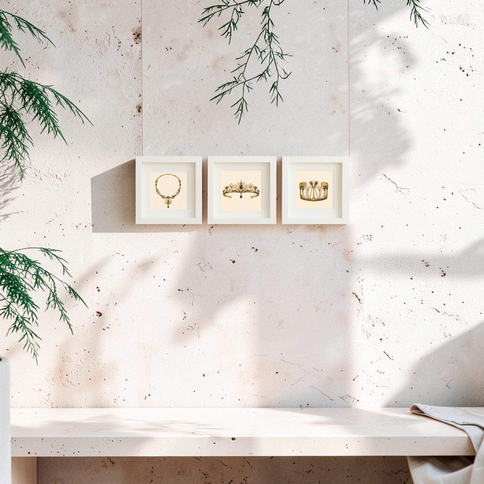 Three framed gold illustrations from Mirabilia Boutique’s “Adornments of the Nile | Mirabilia Relics” collection—a lapis and gold necklace, an ornate crown, and elegant vases inspired by ancient Egyptian jewelry—hang on a light, textured wall above a white bench. Green foliage enters from the left as sunlight casts soft shadows across the Mirabilia canvas artwork, creating a serene atmosphere.