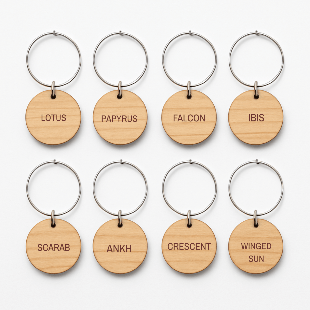 Eight wooden keychains with metal rings, each engraved with a distinct word—LOTUS, PAPYRUS, FALCON, IBIS, SCARAB, ANKH, CRESCENT, and WINGED SUN—are beautifully arranged in two rows. The Ancient Egyptian Wine Charms by Mirabilia Boutique | Lapis & Lotus feature designs inspired by traditional Egyptian motifs, making them the ideal wine accessories to bring a touch of ancient elegance to your gatherings.