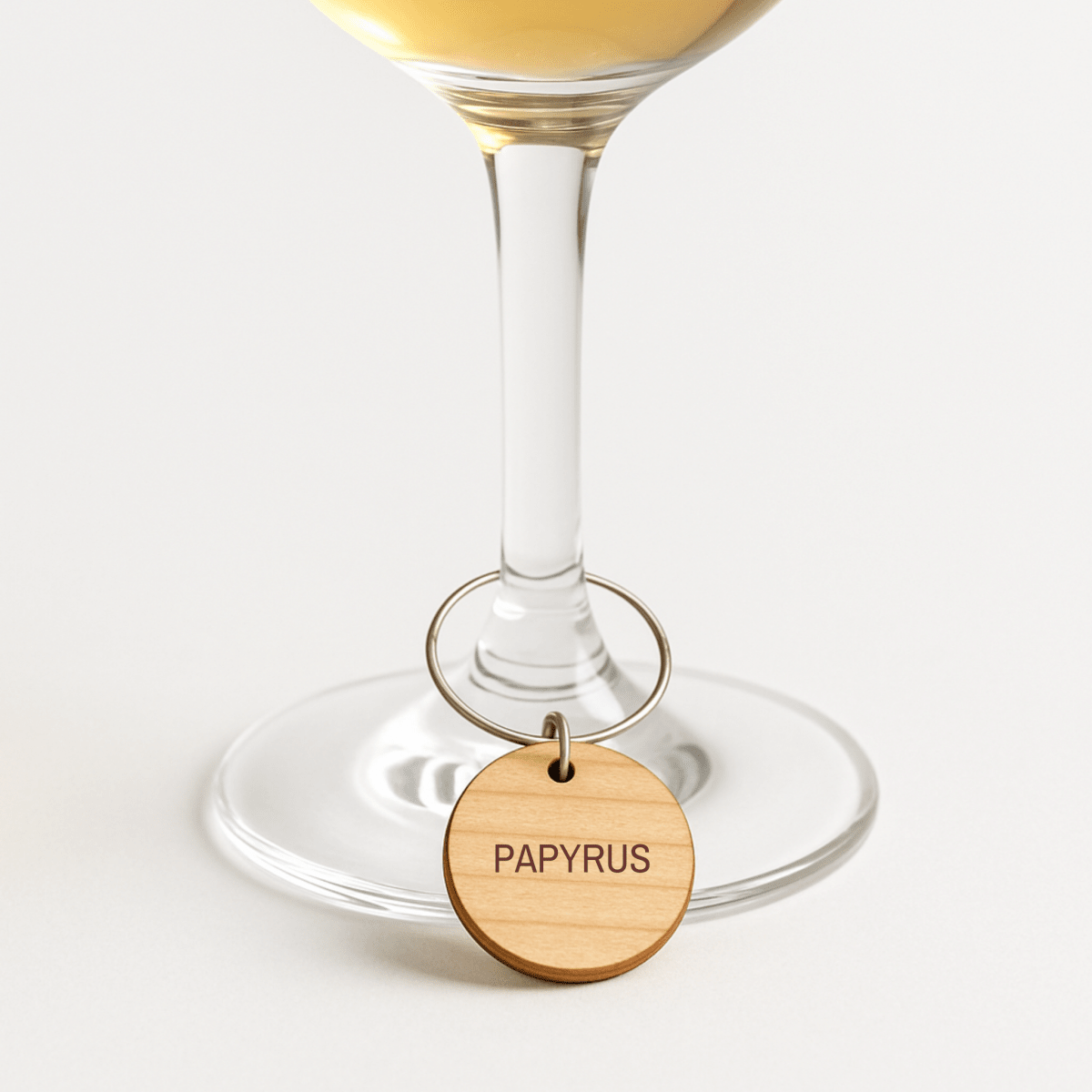 A wooden wine glass charm from the “Ancient Egyptian Wine Charms | Lapis & Lotus” collection by Mirabilia Boutique hangs on the stem of a wine glass filled with white wine, set against a plain white background—an elegant accent that brings a touch of historical charm to any gathering.