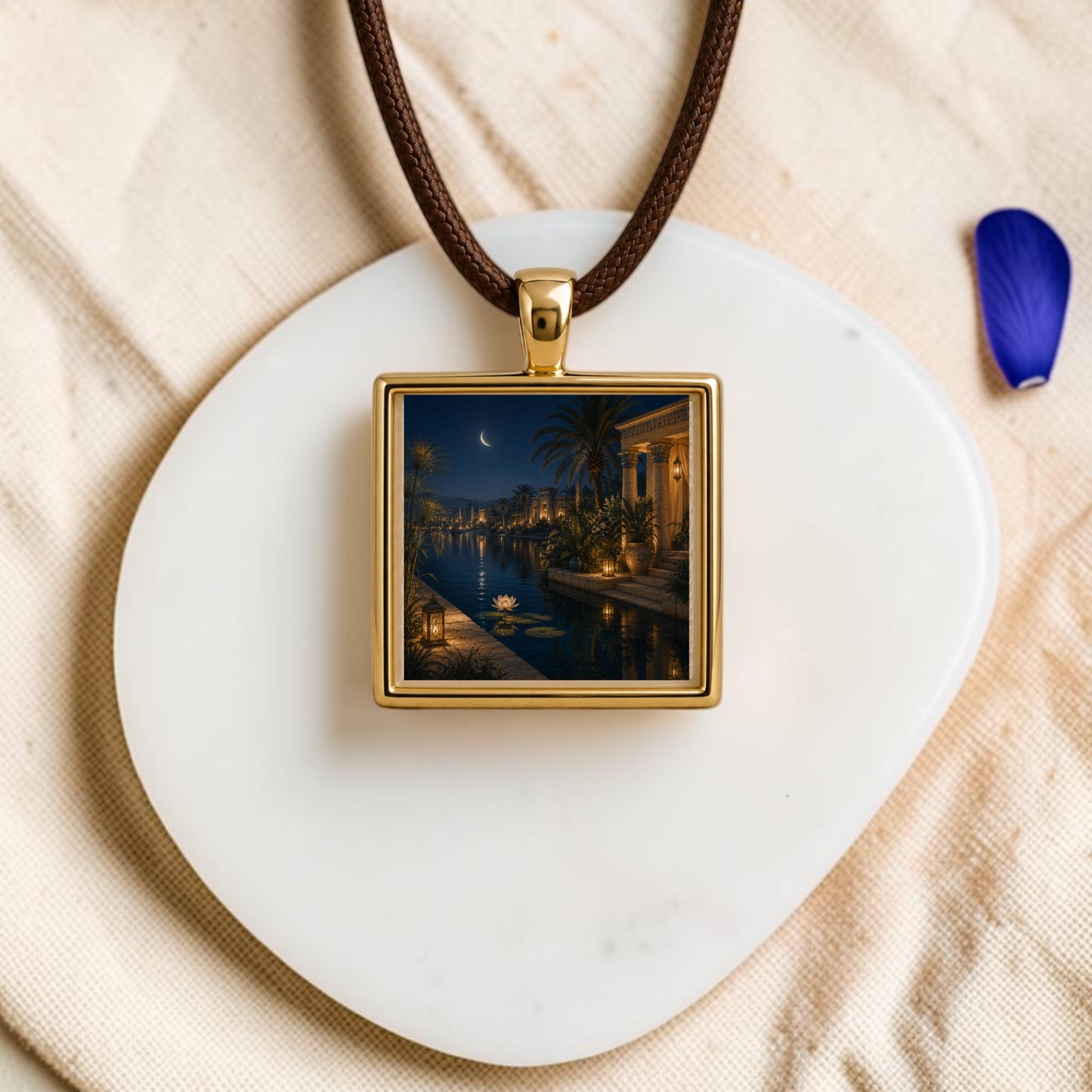 The Lotus After Dark | Scene Pendant Necklace by Mirabilia Boutique features a gold square lotus pendant on a brown cord, resting on a white stone. Showcasing a miniature night scene with palm trees, water lilies, and a crescent moon, this unique piece of wearable art is accented by a single purple petal placed nearby on the beige fabric background.
