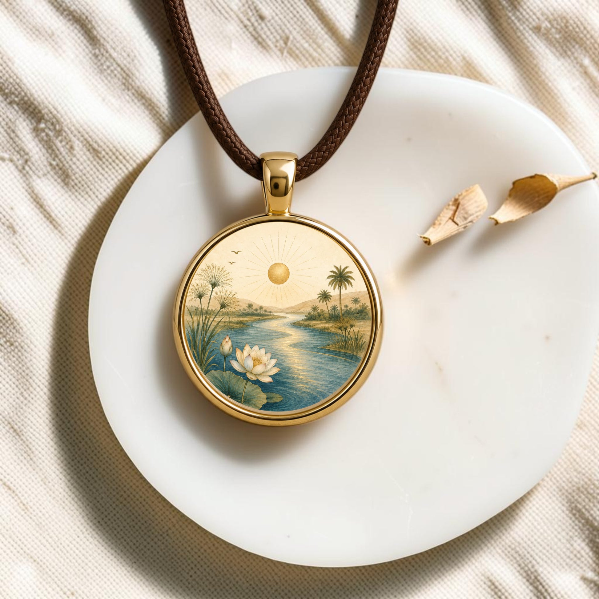 The “Trust the Journey | Story Pendant Necklace” by Mirabilia Boutique—a gold pendant necklace featuring an illustration of a river, lotus flower, palm trees, and sun that captures the essence of Nile landscape jewelry as a stunning piece of wearable art—rests on a white stone with two dried flower petals nearby.