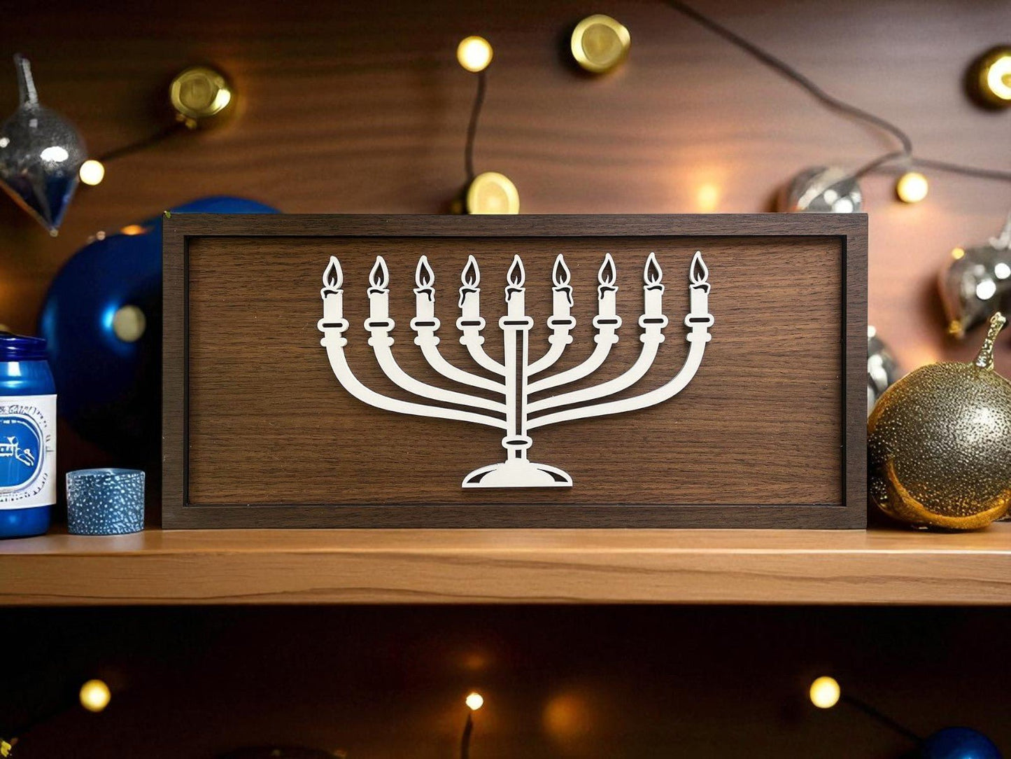 The Mirabilia Boutique 18x8 Modern Menorah Sign features a minimalist white laser-cut menorah in a wooden frame, displayed on a shelf with Hanukkah decor such as blue and gold ornaments, candles, and string lights.