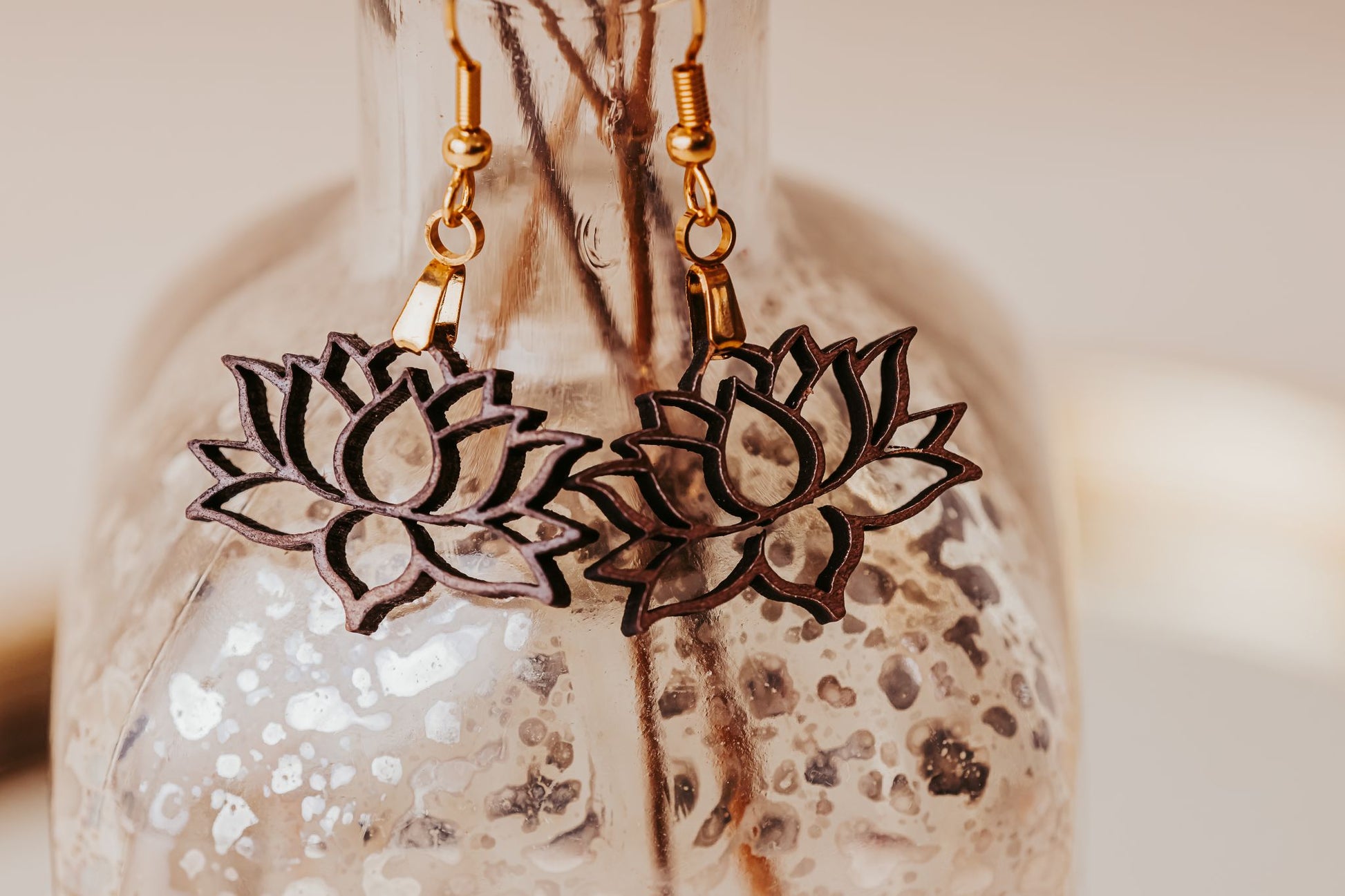 A close-up of Mirabilia Boutique’s Laser Cut Lotus Flower Wood Earrings—lightweight, boho-style pieces with gold hooks—hanging on a textured glass bottle, showcasing their delicate outlined design.