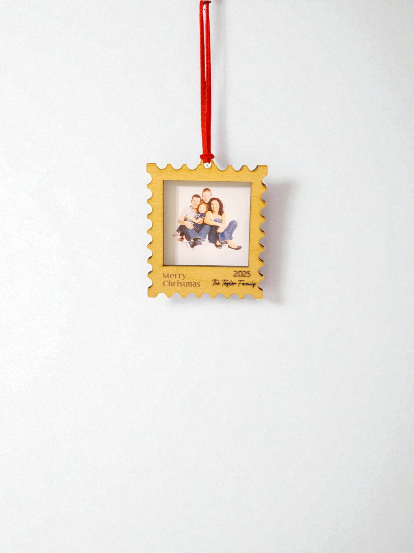 The Mirabilia Boutique Personalized Christmas Stamp Frame Ornament hangs from a red ribbon, showcasing a family photo. “Merry Christmas 2025” and “The Taylor Family” are inscribed on its white background—a custom gift for your loved ones.