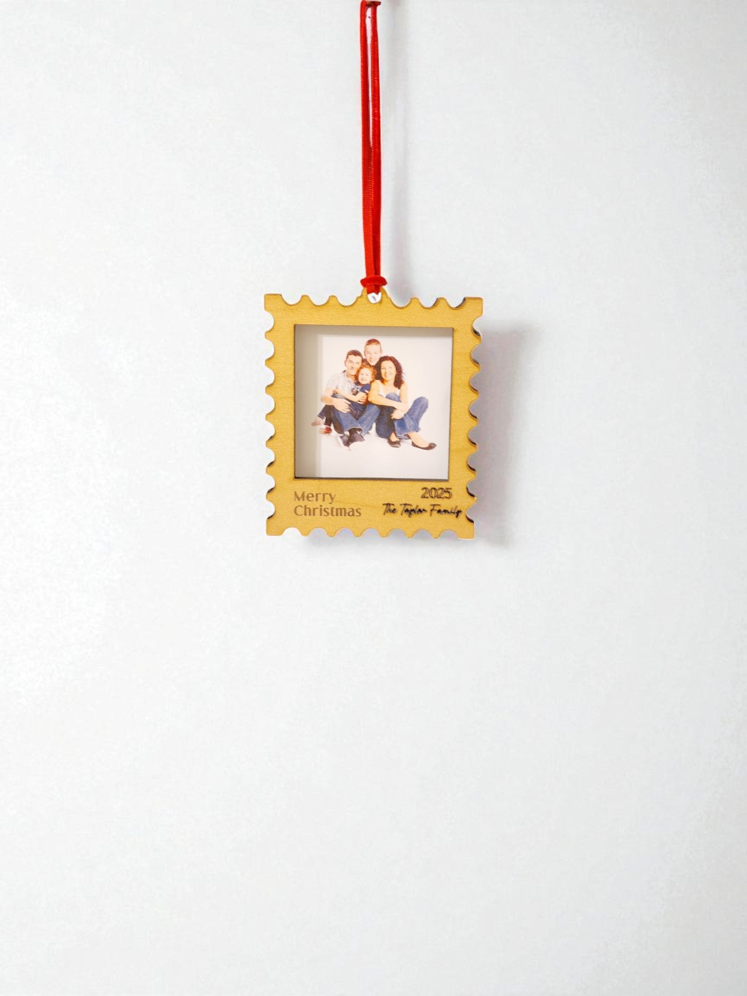 The Mirabilia Boutique Personalized Christmas Stamp Frame Ornament hangs from a red ribbon, showcasing a family photo. “Merry Christmas 2025” and “The Taylor Family” are inscribed on its white background—a custom gift for your loved ones.