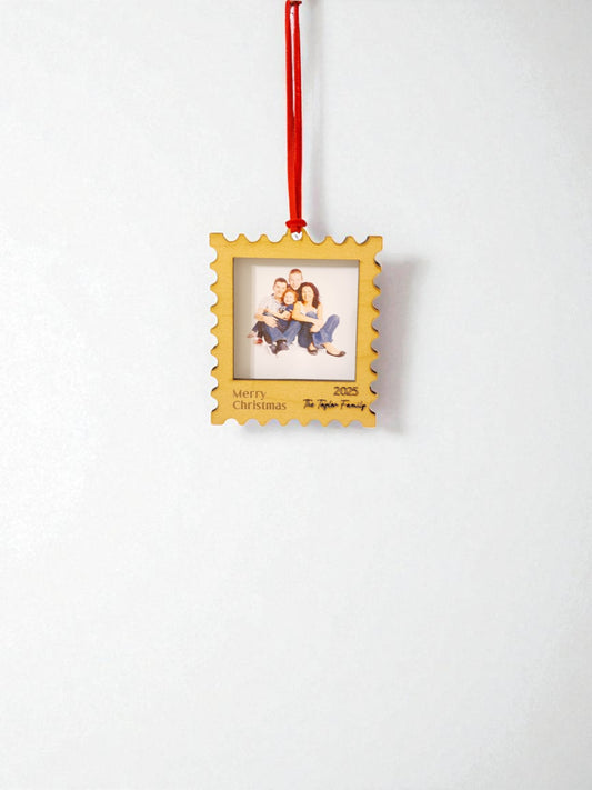 The Mirabilia Boutique Personalized Christmas Stamp Frame Ornament hangs from a red ribbon, showcasing a family photo. “Merry Christmas 2025” and “The Taylor Family” are inscribed on its white background—a custom gift for your loved ones.