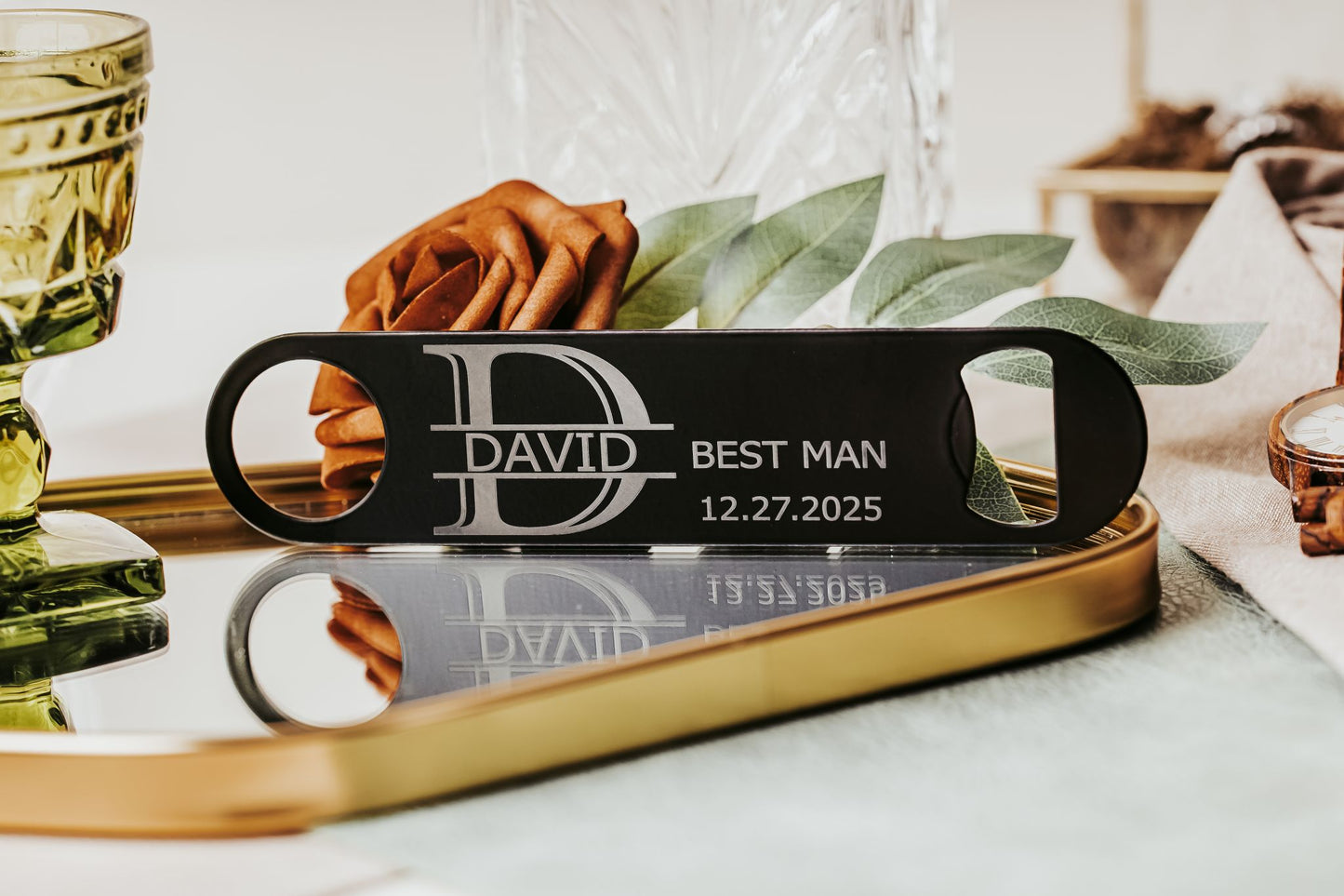 The Engraved Flat Metal Bottle Opener by Mirabilia Boutique, featuring "David," "Best Man," and "12.27.2025," sits on a mirrored tray with leaves and glassware—an ideal groomsmen gift for your special day.