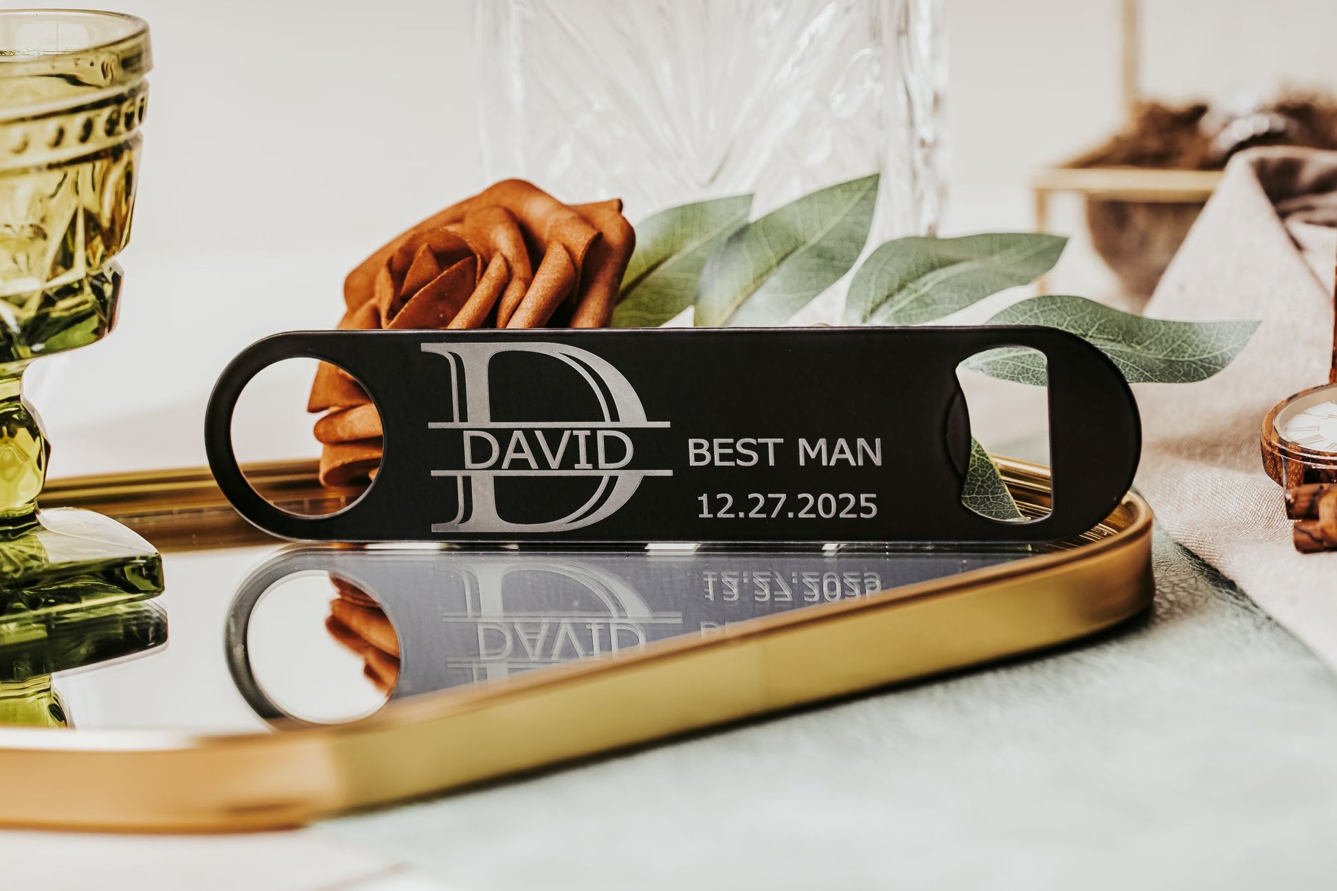 The Engraved Flat Metal Bottle Opener by Mirabilia Boutique, featuring "David," "Best Man," and "12.27.2025," sits on a mirrored tray with leaves and glassware—an ideal groomsmen gift for your special day.