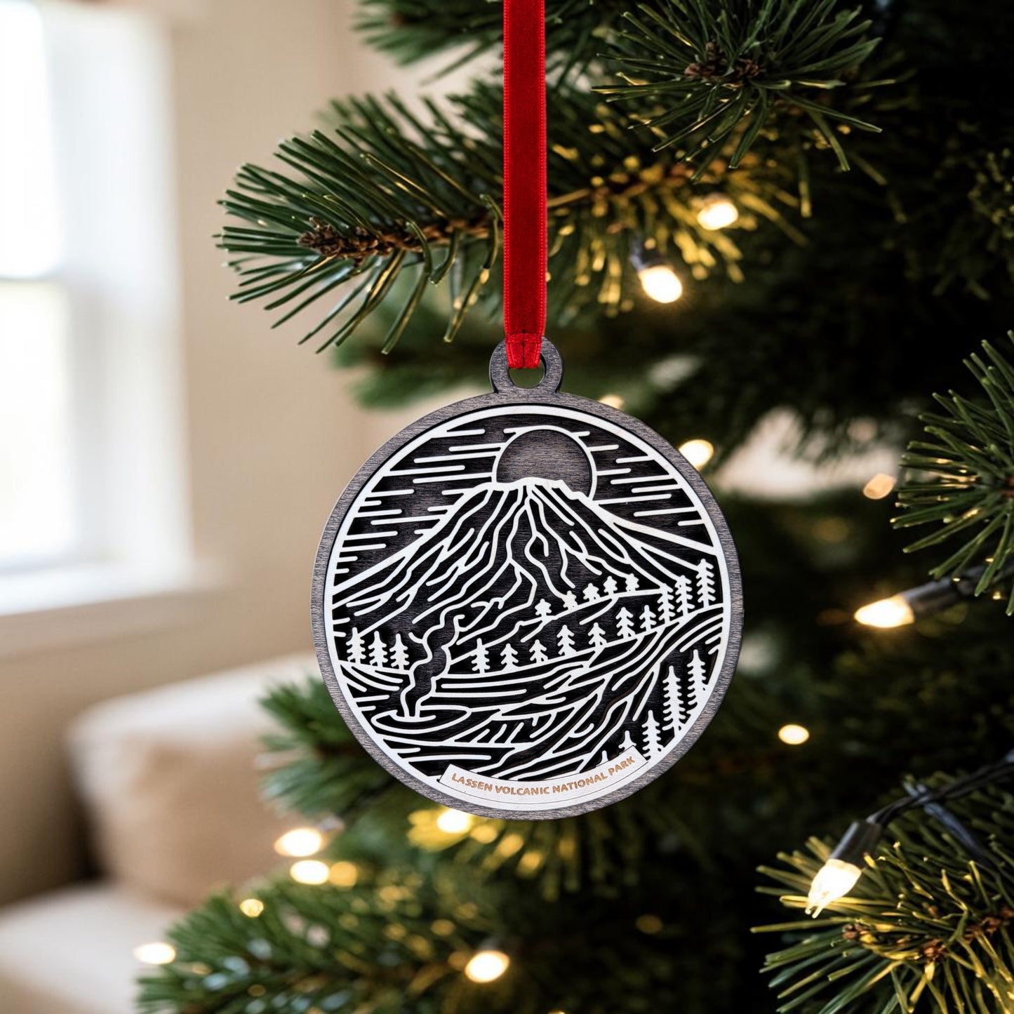 The Lassen Volcanic National Park Line Art Christmas Ornament by Mirabilia Boutique, featuring a mountain and tree design inspired by Lassen Peak, hangs from a Christmas tree branch with a red ribbon amid glowing lights.